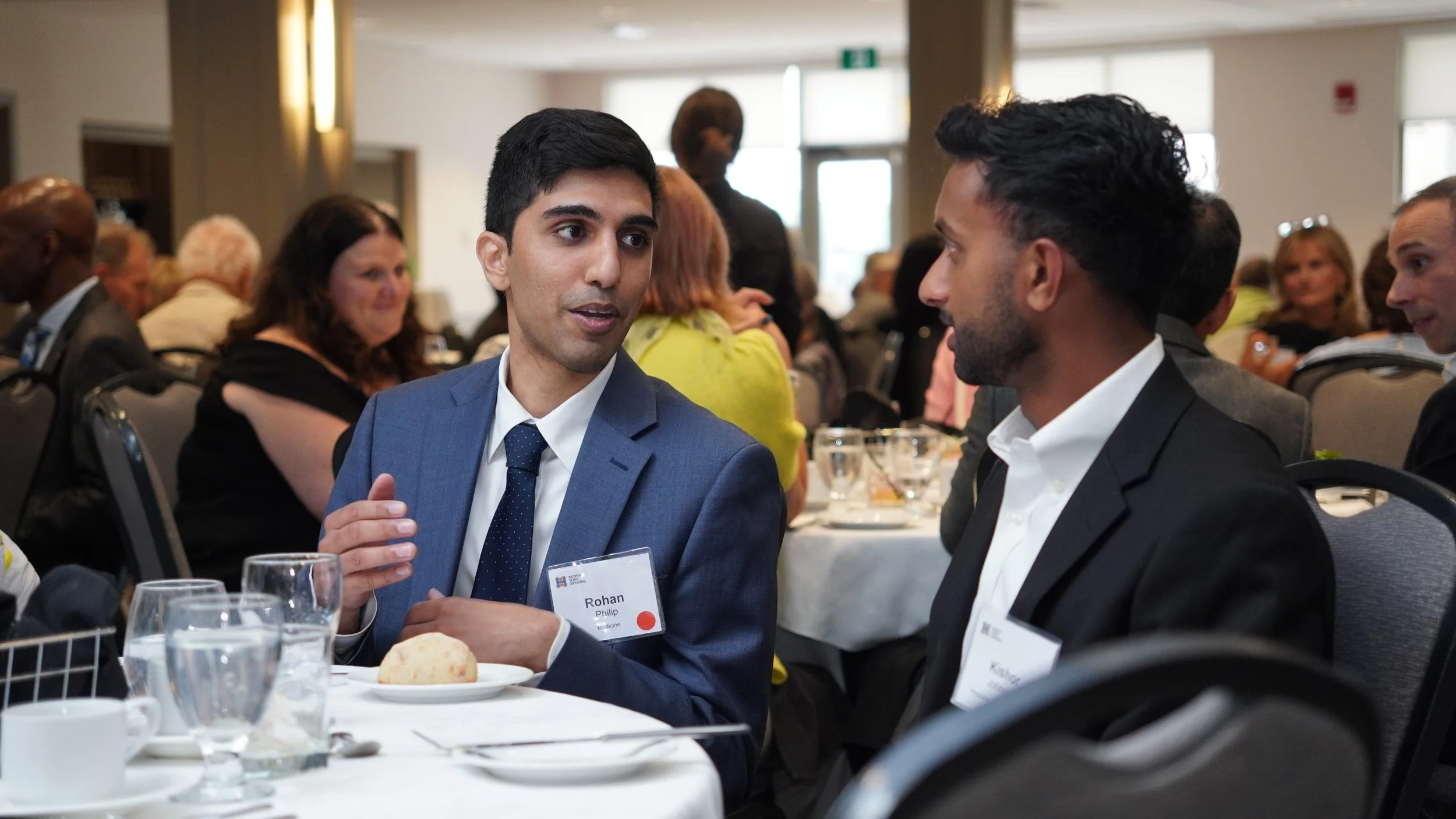 Two men in suits with name tags engaged in conversation at a banquet table in a conference room with other attendees in the background.