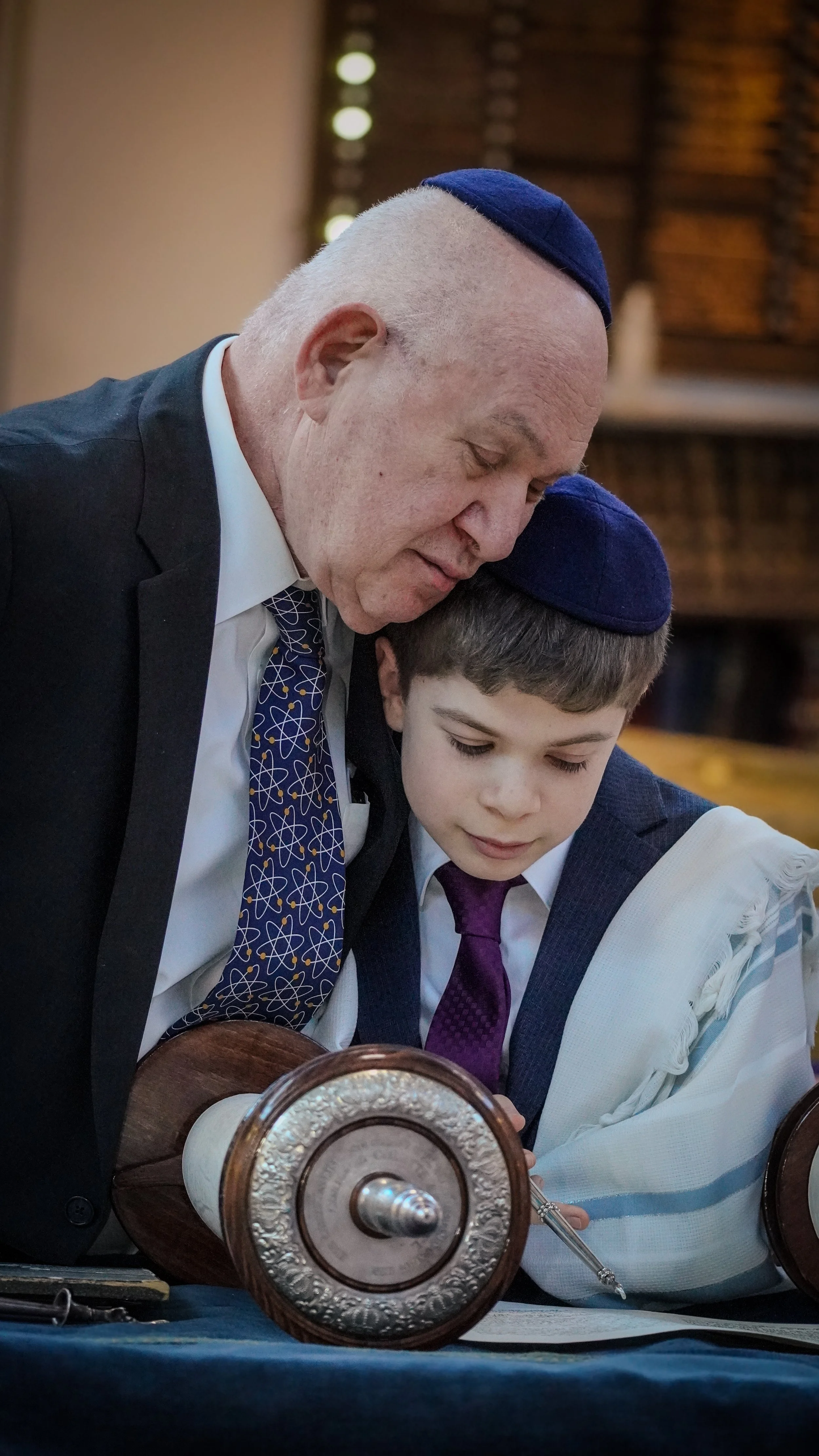 An older man and a young boy, both wearing kippahs, are leaning over a table quietly reading or studying a document together.