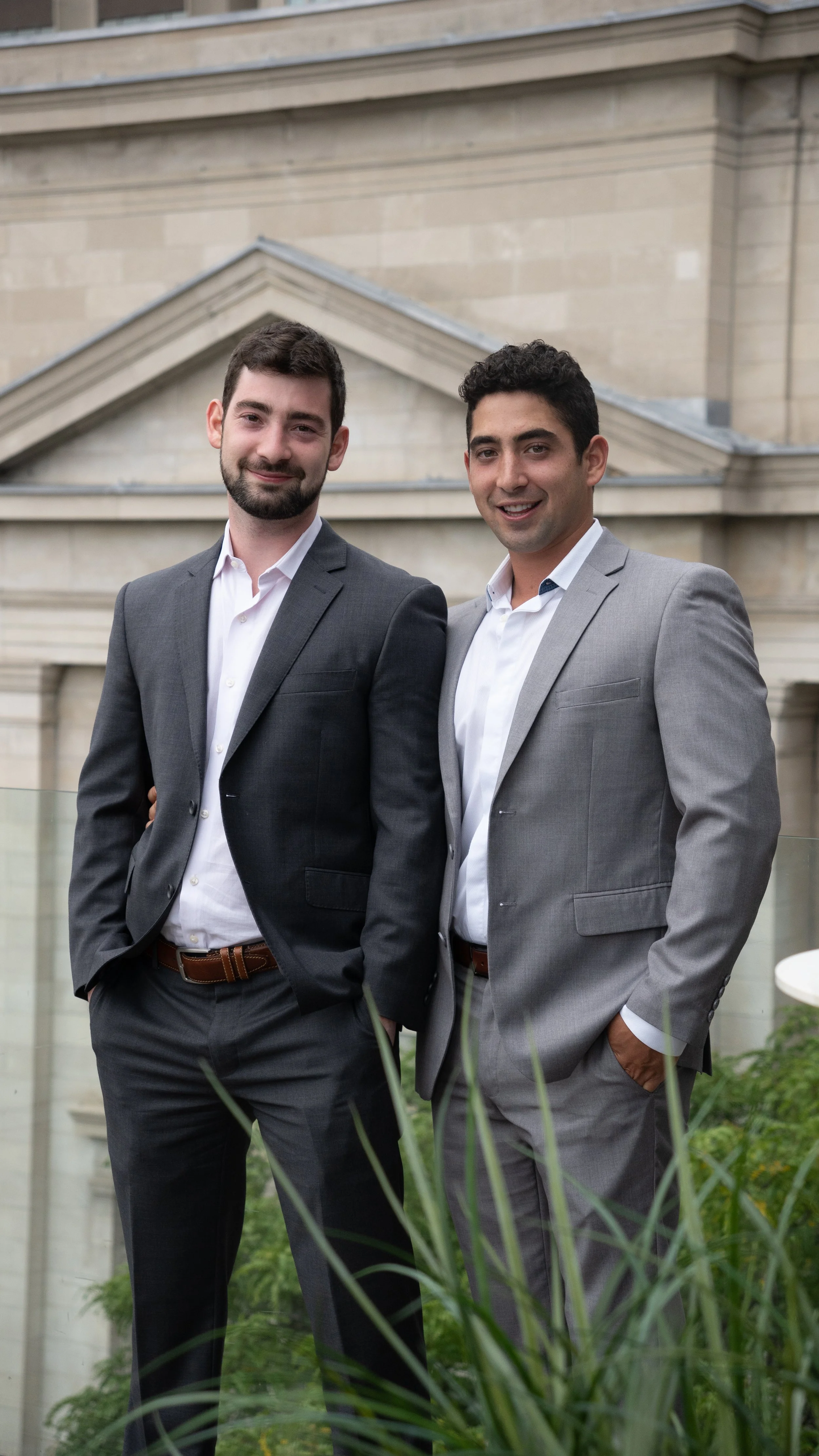 Two men in suits standing outdoors in front of a building with classical architecture, smiling at the camera.