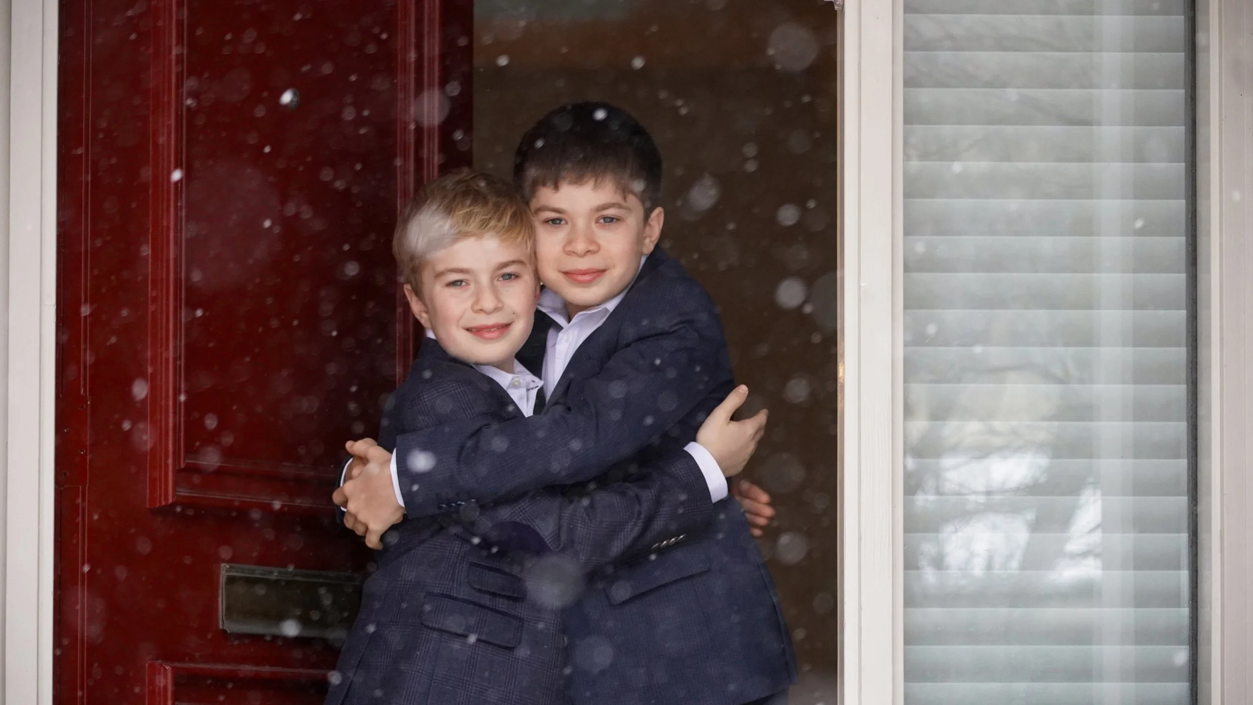 Two boys in suits hugging and smiling at the camera in front of a red door during snowfall.