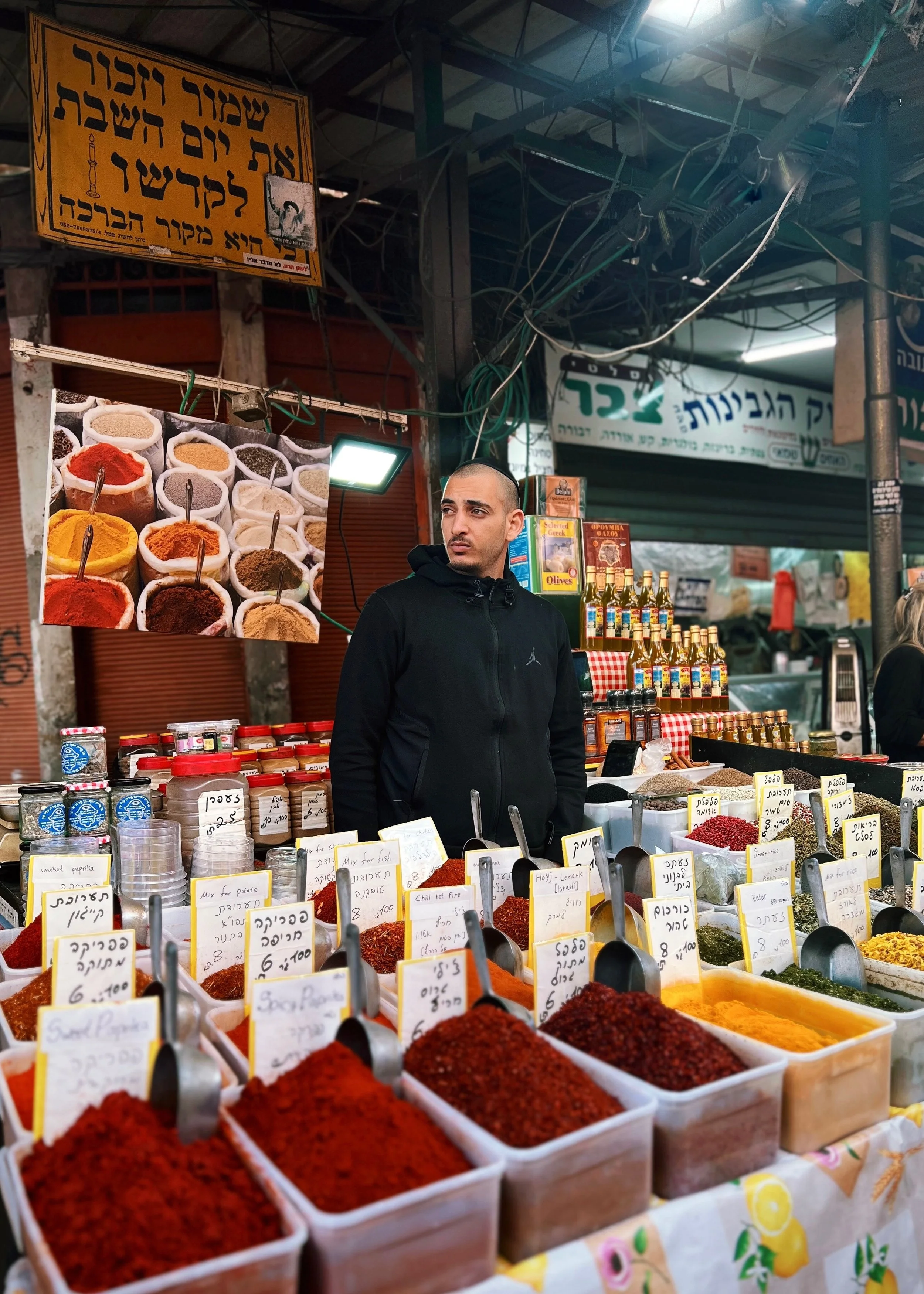 A man in a black hoodie stands behind a stall selling various colorful spices and herbs at an outdoor market. The spices are in white containers with small scoops, and there are handwritten signs in Hebrew indicating names and prices.