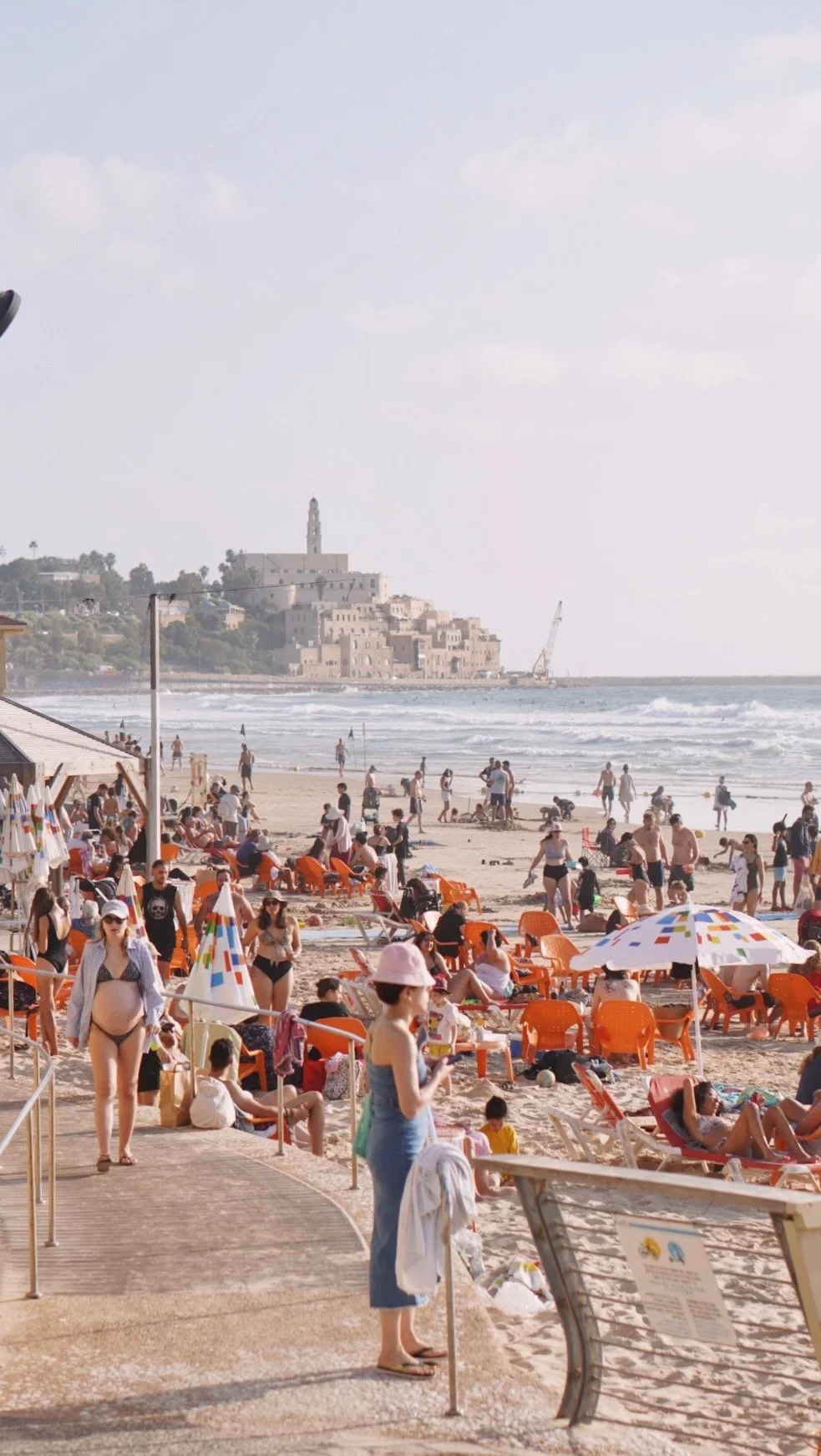 Beach scene with people sunbathing, walking, and playing near the shore; orange lounge chairs, umbrellas, and a sandy beach with a city and hillside in the background.