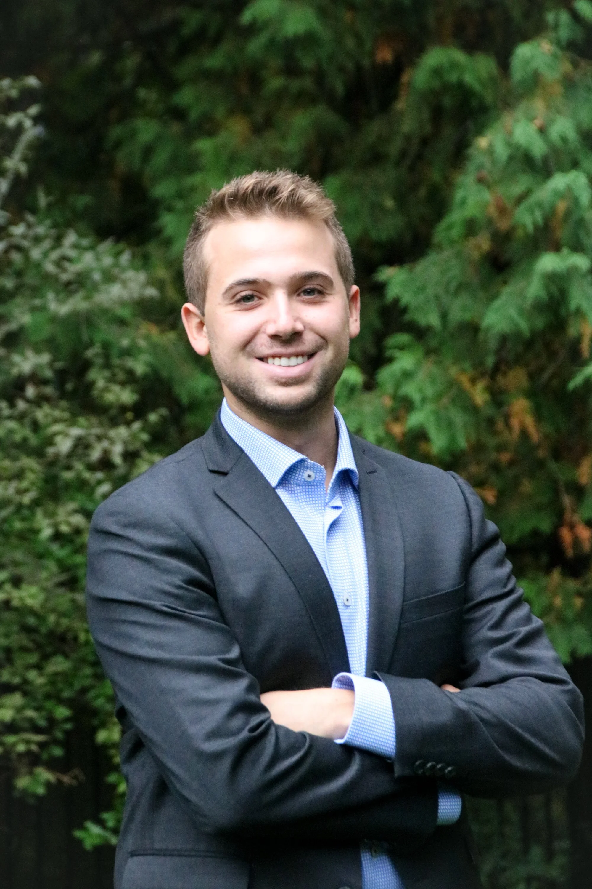 Portrait of a young man in a suit with crossed arms, smiling, outdoors with green foliage in the background.