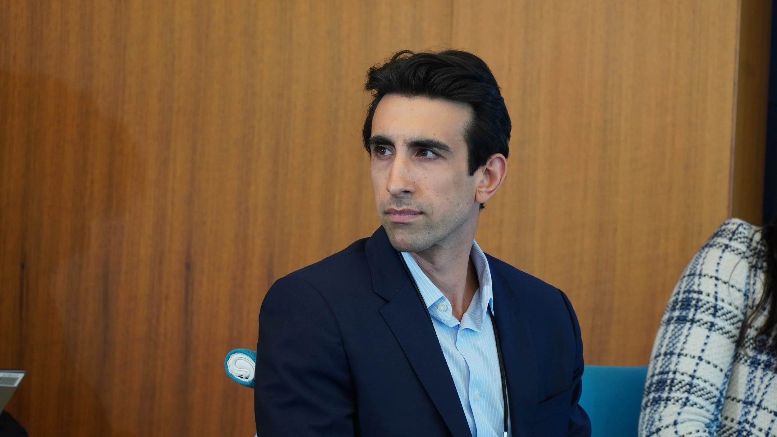A man with dark hair and a clean-shaven face wearing a navy blazer and light blue shirt looking to his left, sitting in a room with wooden paneling in the background.