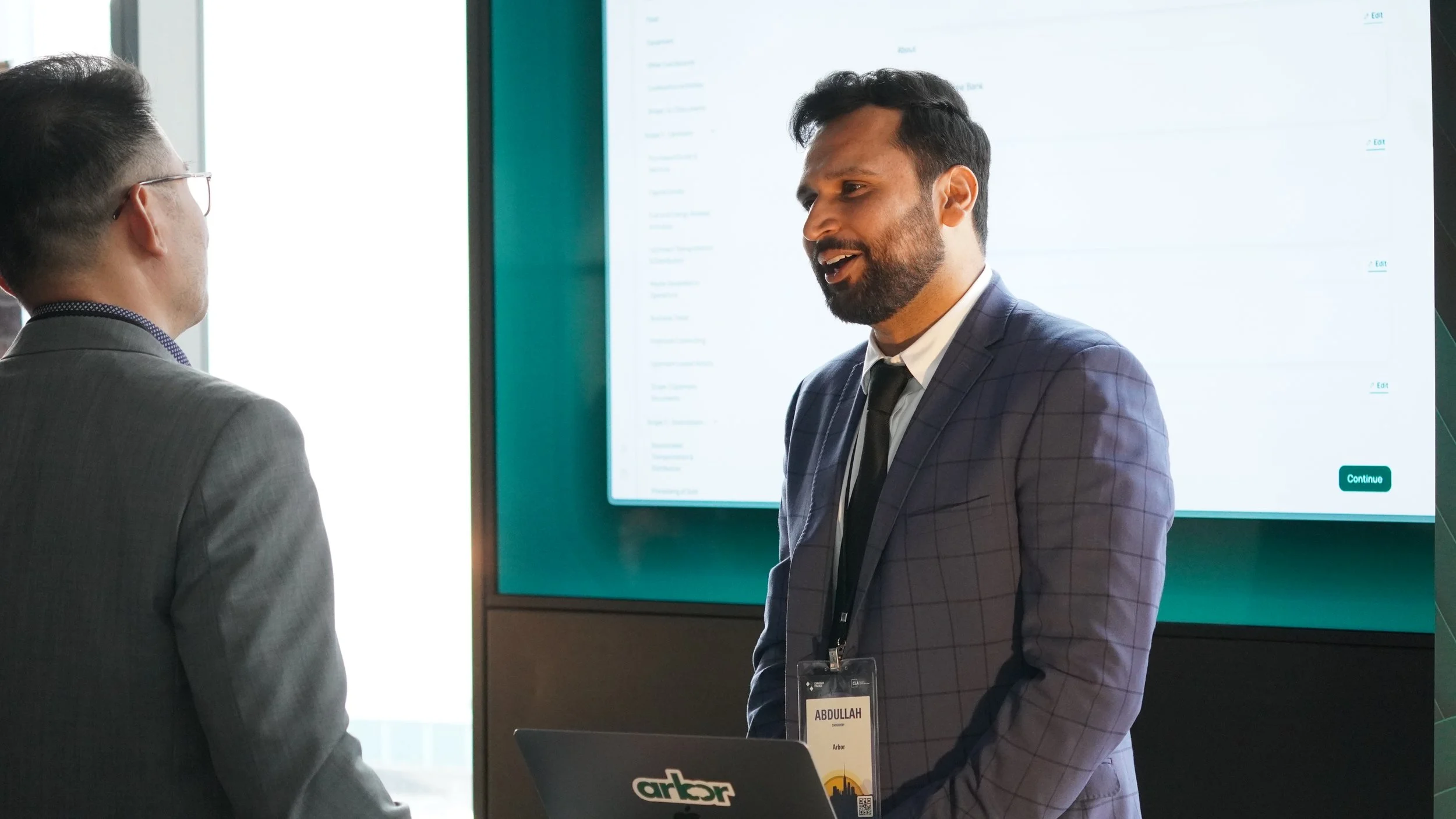 Two men in suits having a conversation at a professional event, with a large presentation screen displaying a dashboard in the background.