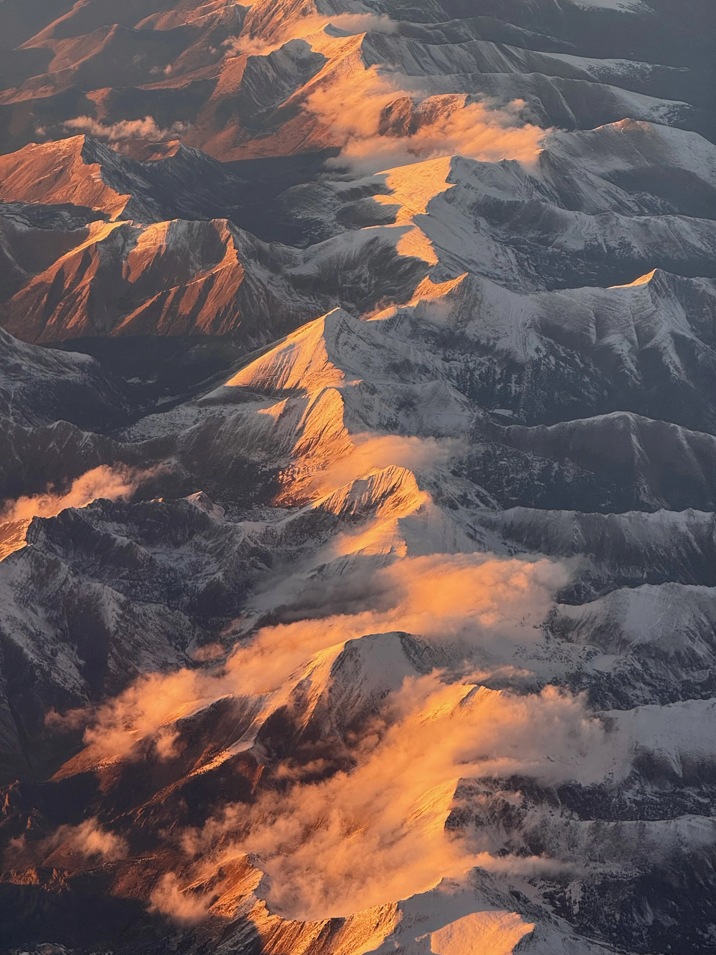 Aerial view of snow-capped mountains during sunset with orange and golden light reflecting on the peaks.