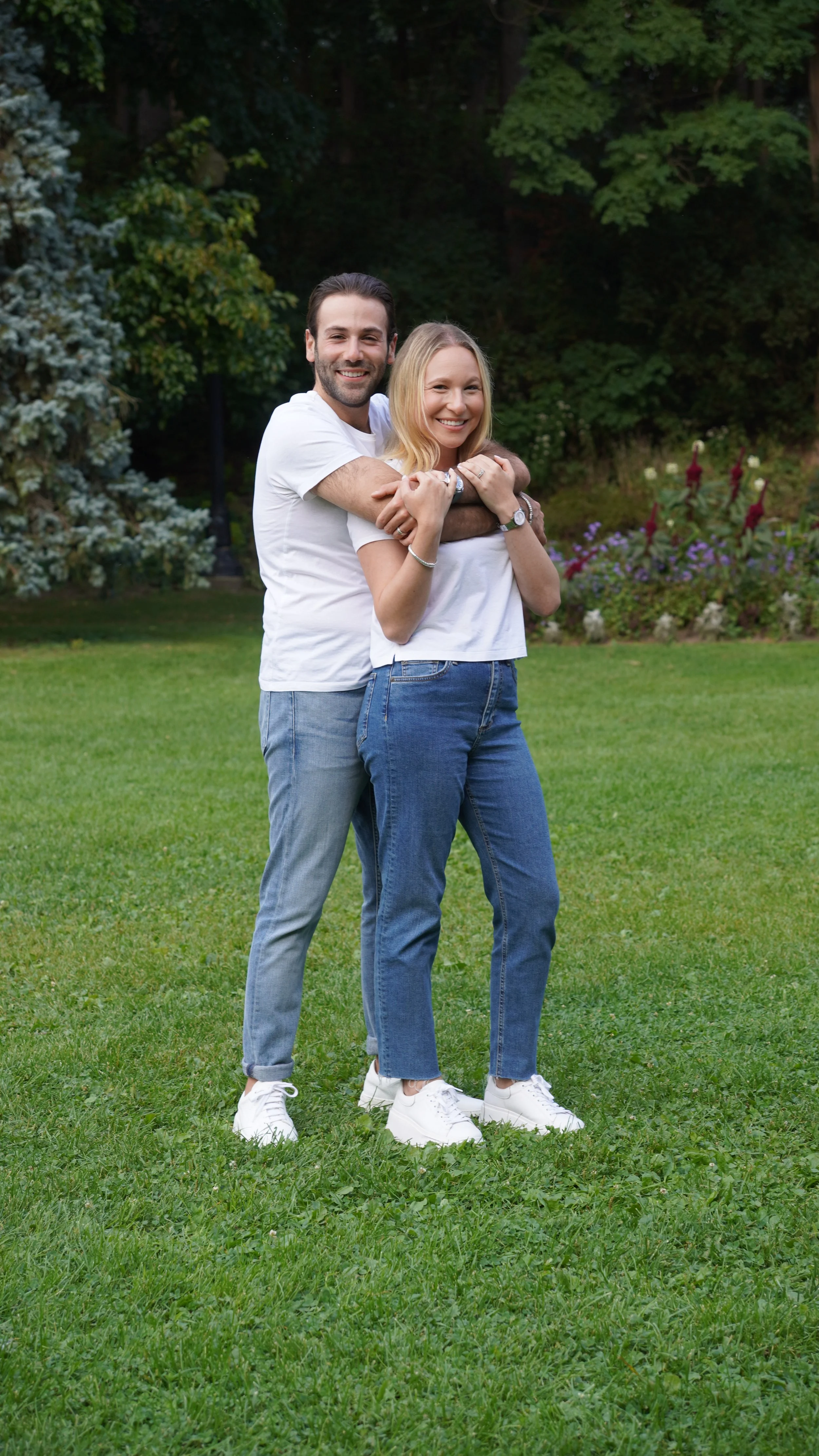 A happy couple stands on grass in a park, with the man hugging the woman from behind. Both are smiling and wearing casual white T-shirts and jeans, with the woman in white sneakers. The background features trees and colorful flowers.