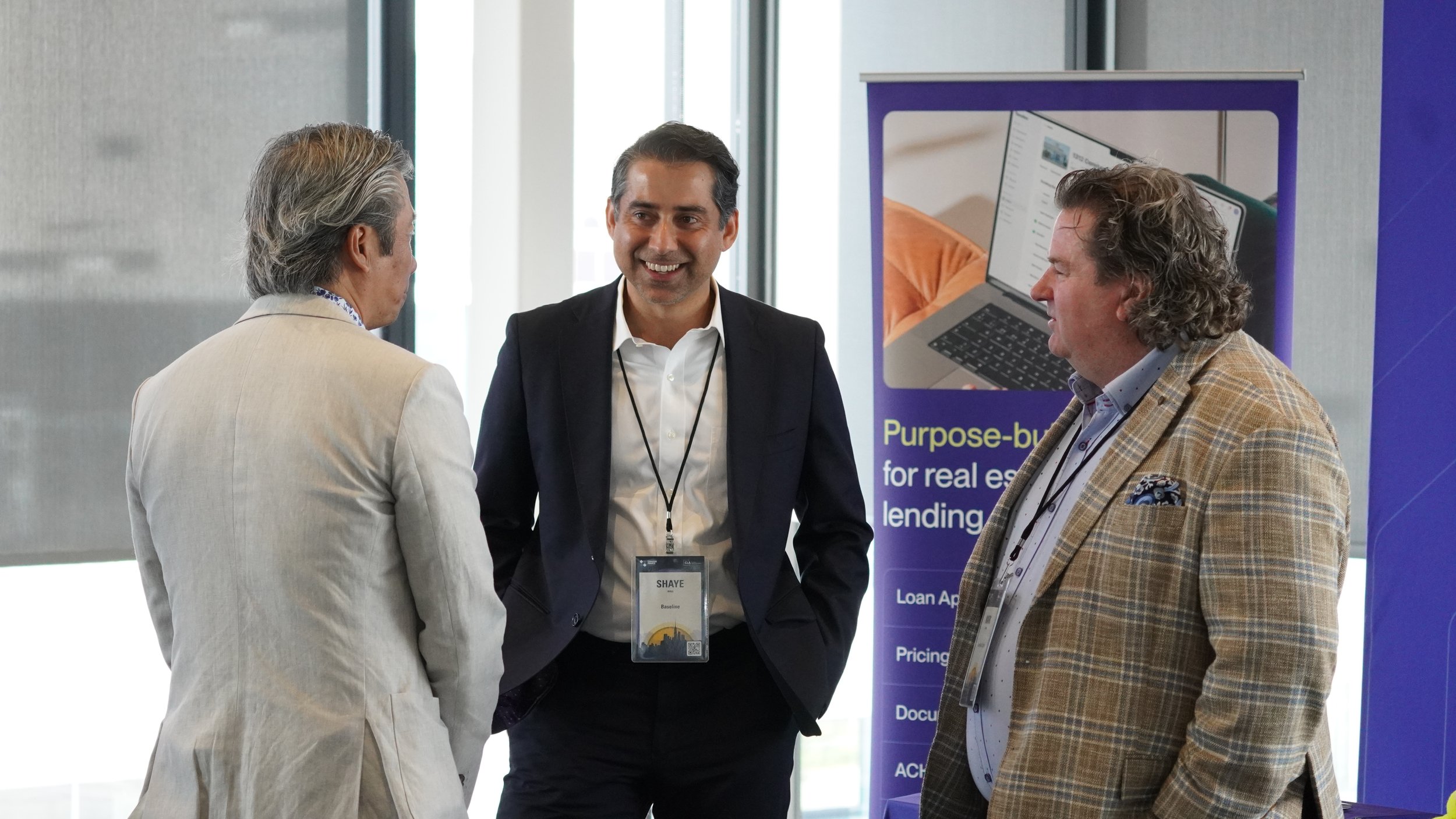 Three men in business attire engaging in conversation at a professional event, with a purple banner mentioning purpose-built solutions for real estate lending in the background.