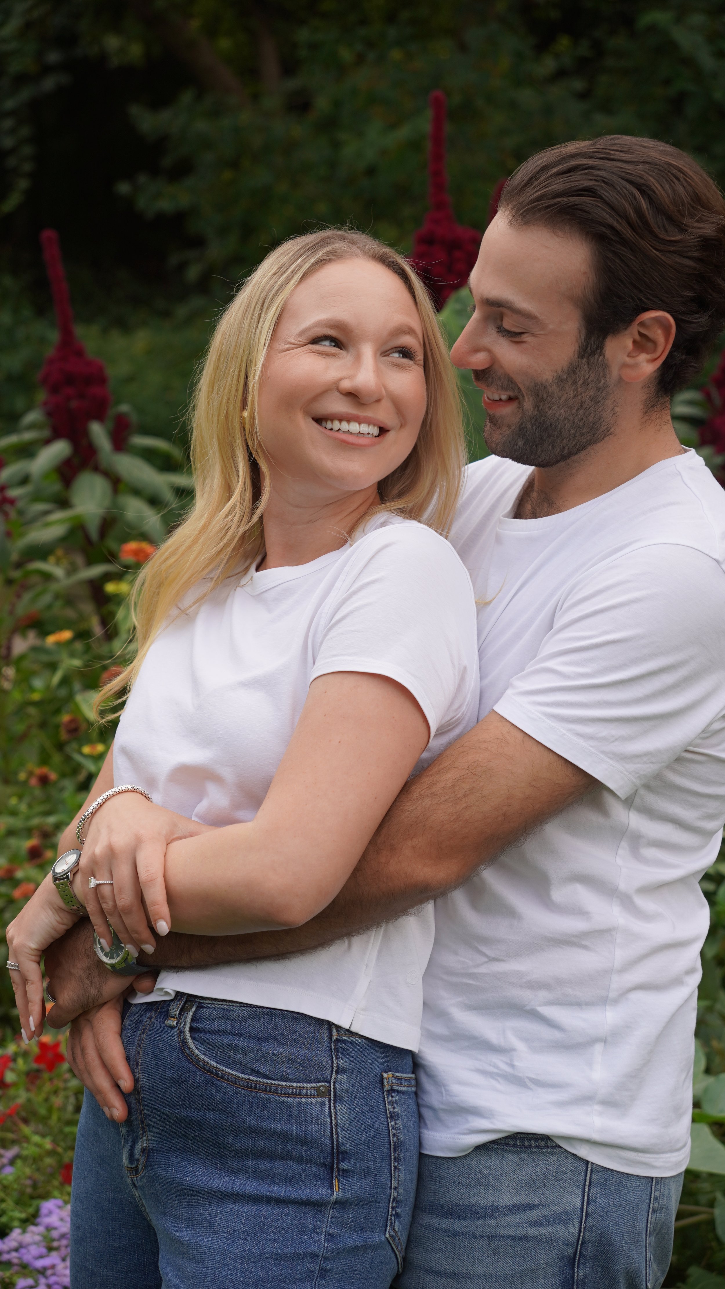 A smiling couple embracing outdoors in a garden with colorful flowers and greenery.