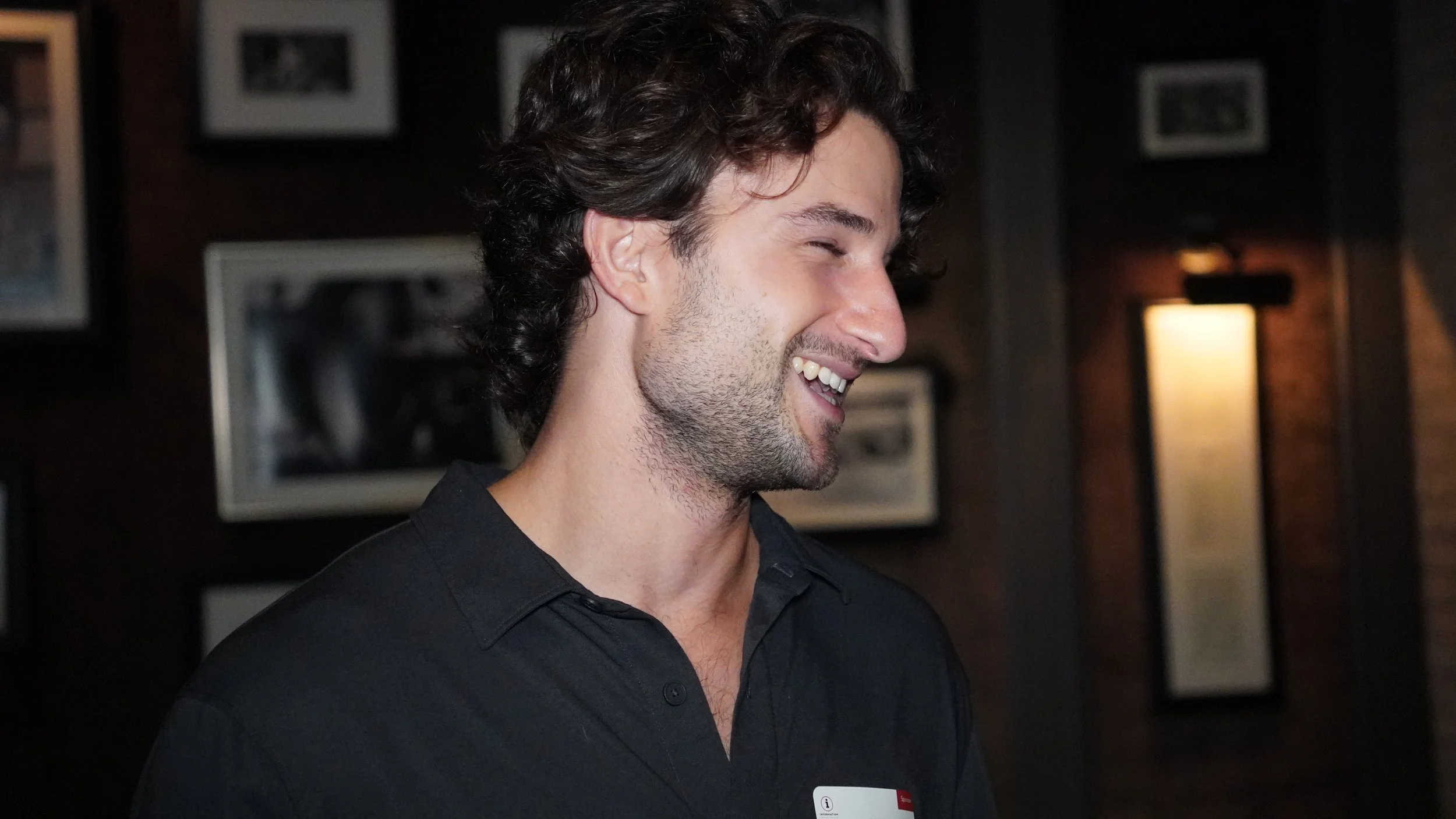 Smiling young man with dark, curly hair wearing a black collared shirt, standing indoors with framed pictures and warm lighting on the dark wall behind him.