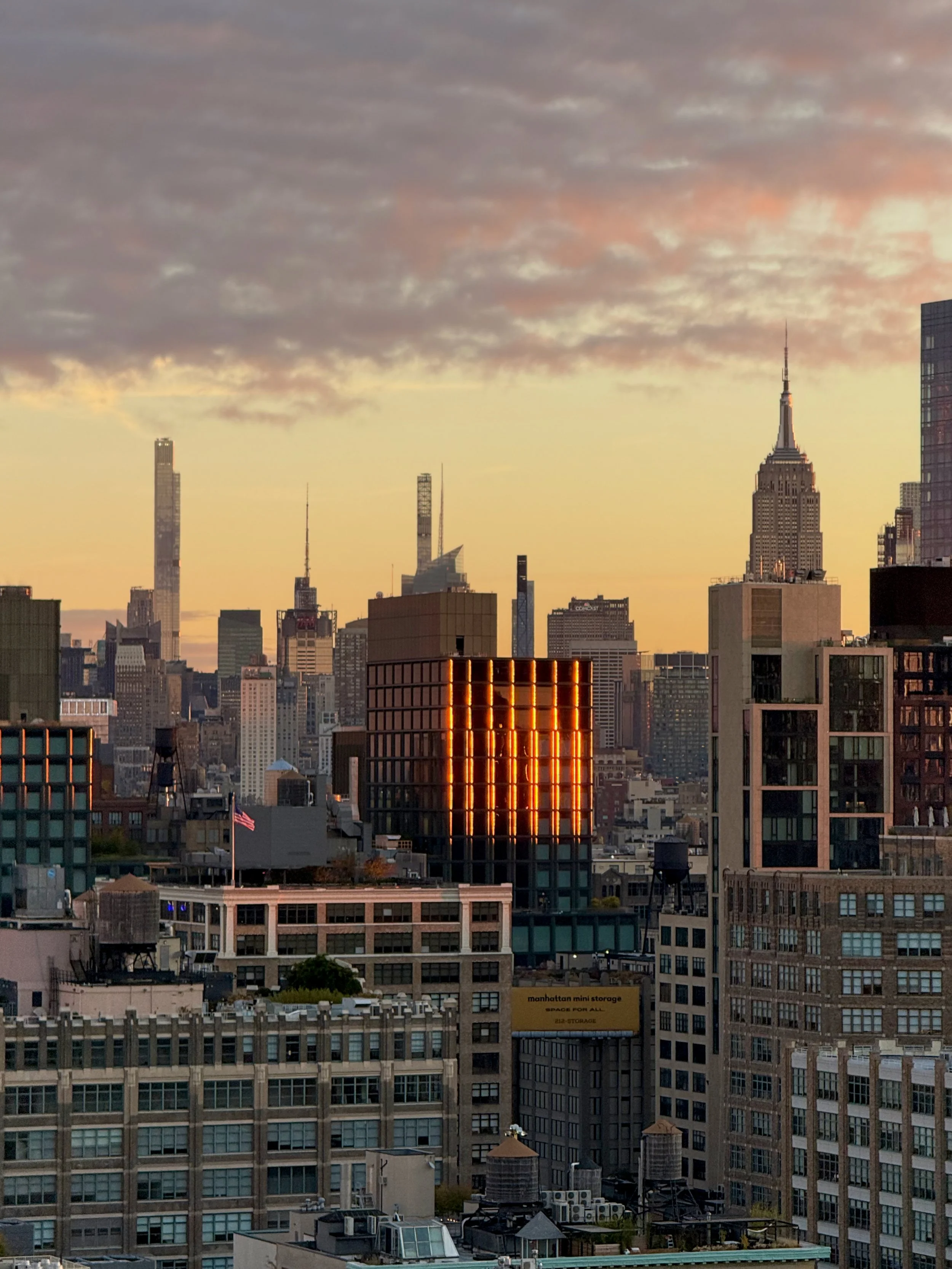 City skyline at sunset with tall buildings, including the Empire State Building and a neon-lit building in the foreground.