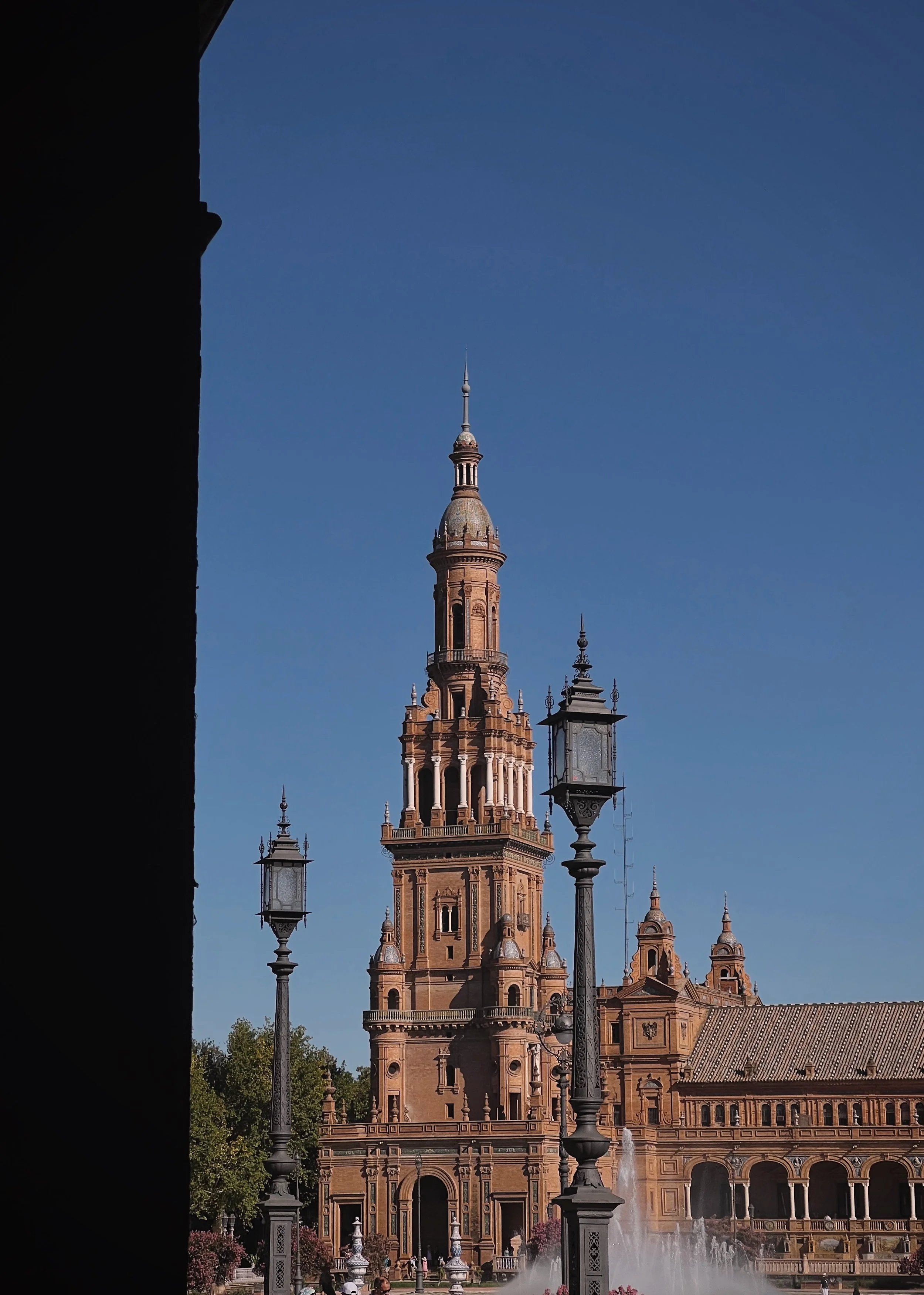 View of a historic building with a tall clocktower and ornate architecture, flanked by antique street lamps, under a clear blue sky.