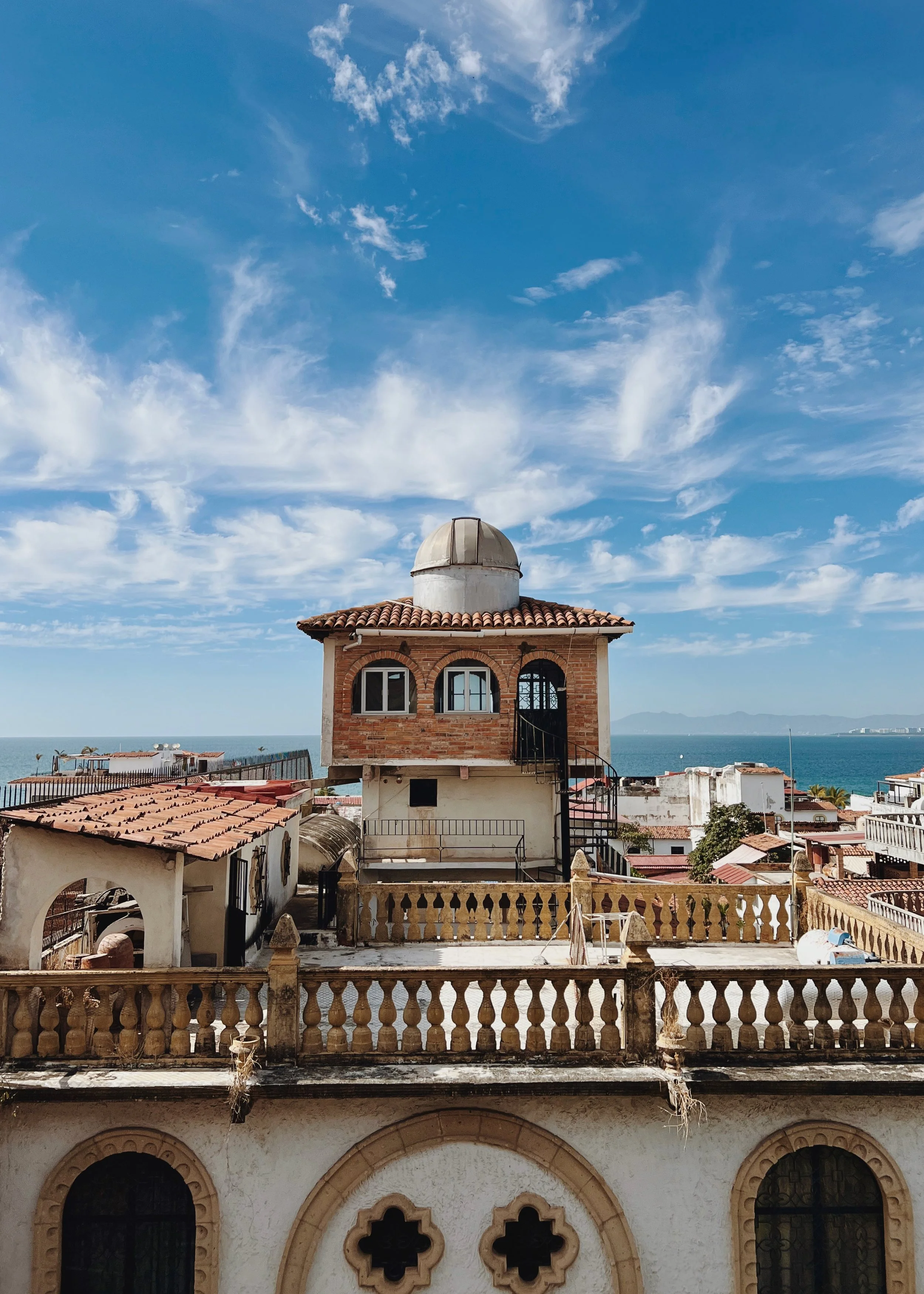 A rooftop view of a house with a brick facade, dome-shaped observatory, and stairway leading to the upper floor, with the sea and sky in the background.
