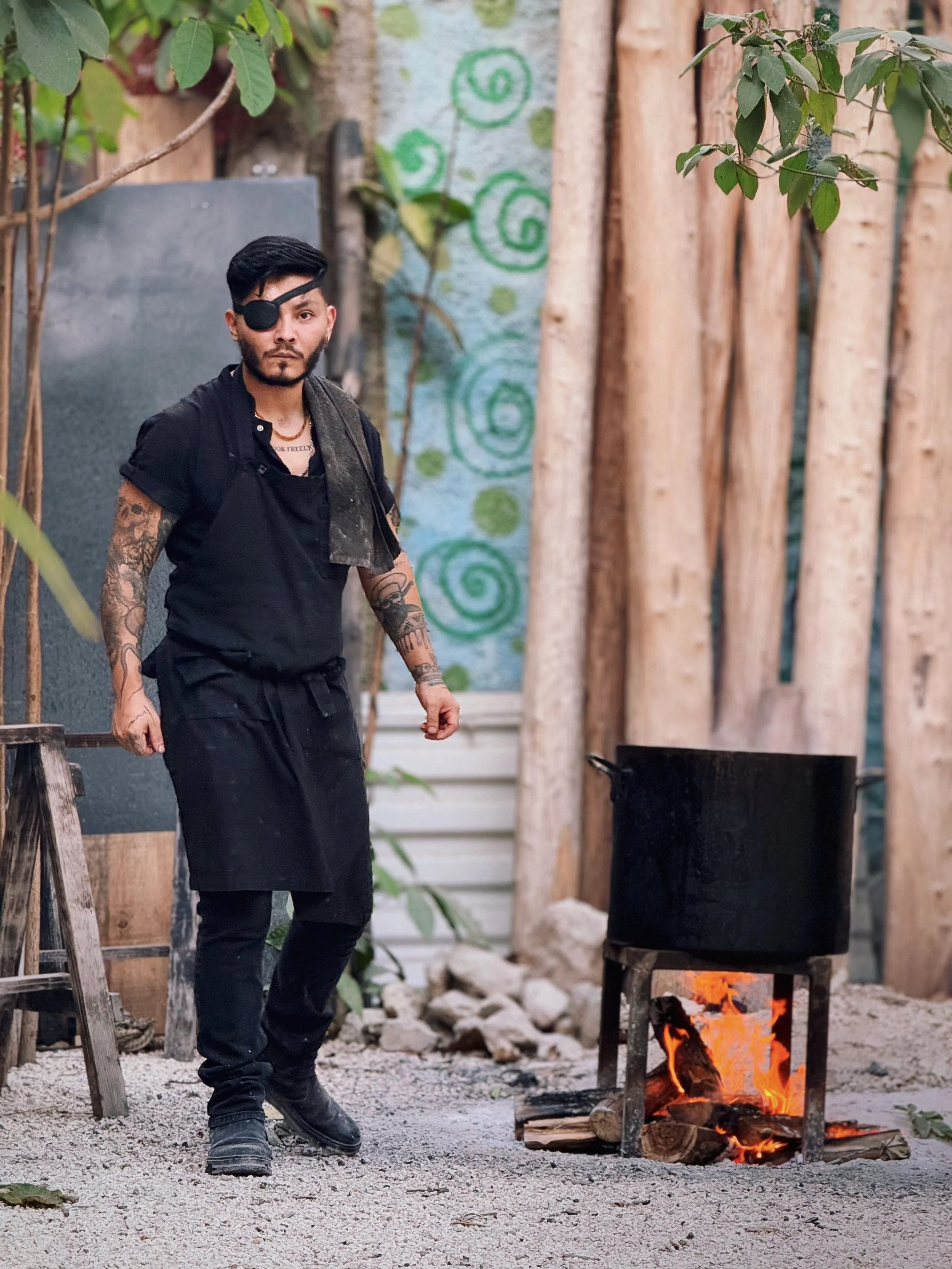 A man with tattoos wearing a black apron and an eye patch standing outdoors near a cooking pot over an open fire, with a wooden fence and decorative green swirls on a wall behind him.
