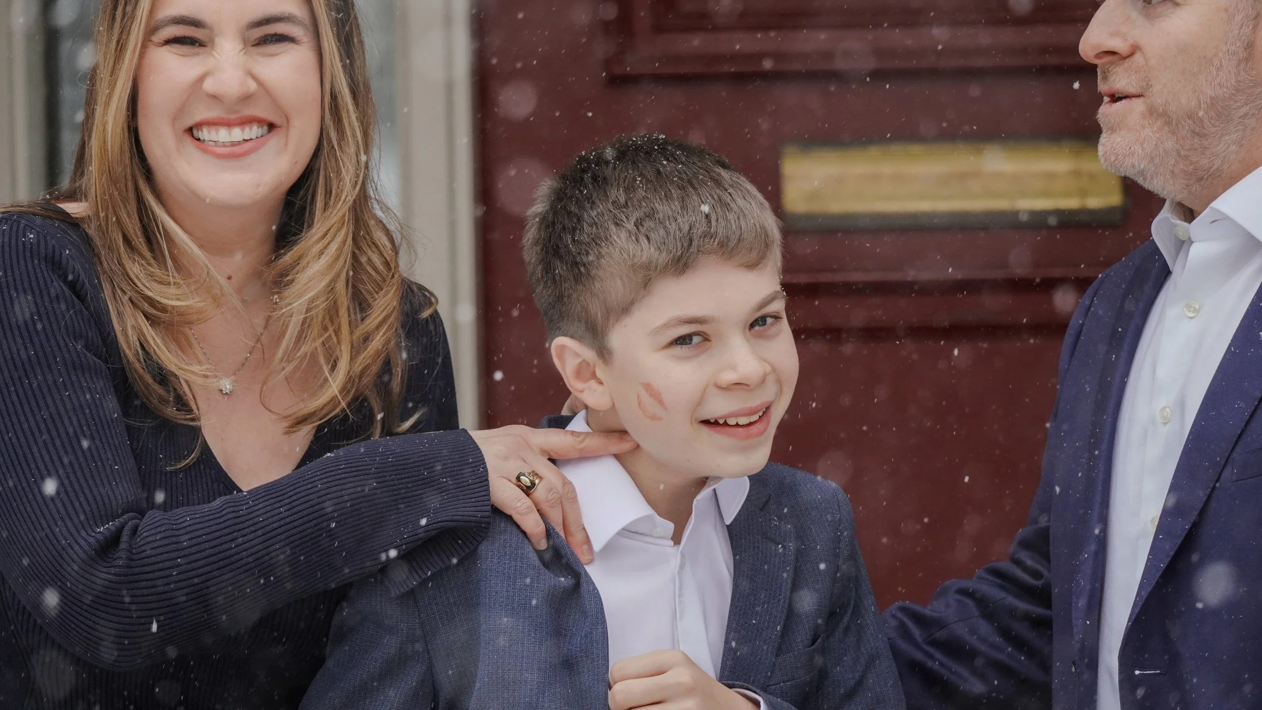 A young boy with a lipstick kiss mark on his face, smiling, being patted on the shoulder by a woman and a man.