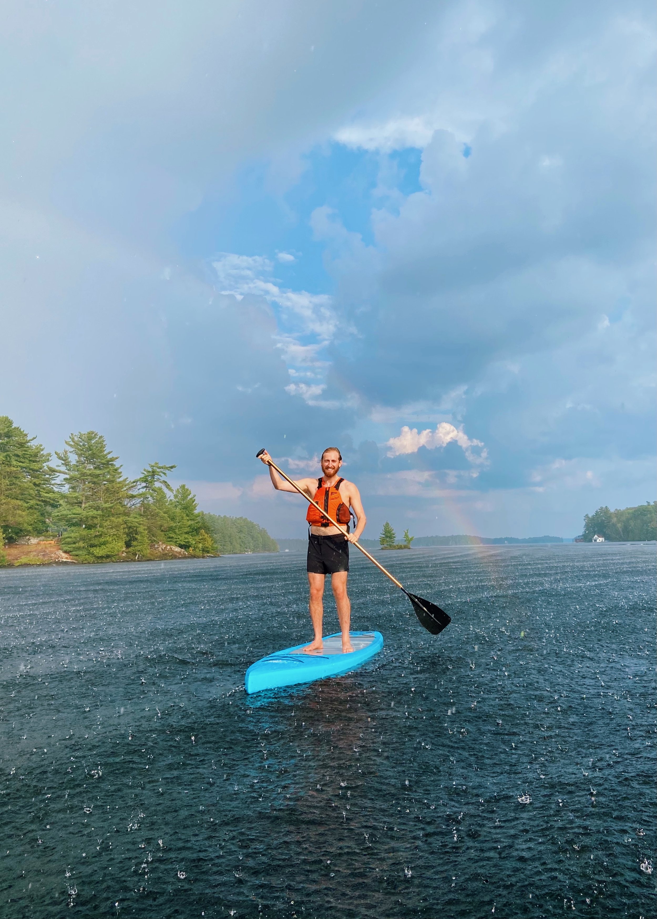 A man paddleboarding on a lake during a rainstorm with a rainbow in the background.