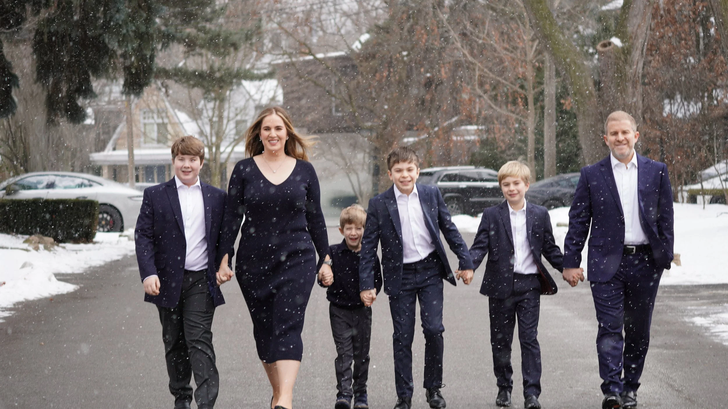 A family of seven walking on a snowy street, holding hands and smiling, with cars and houses in the background.