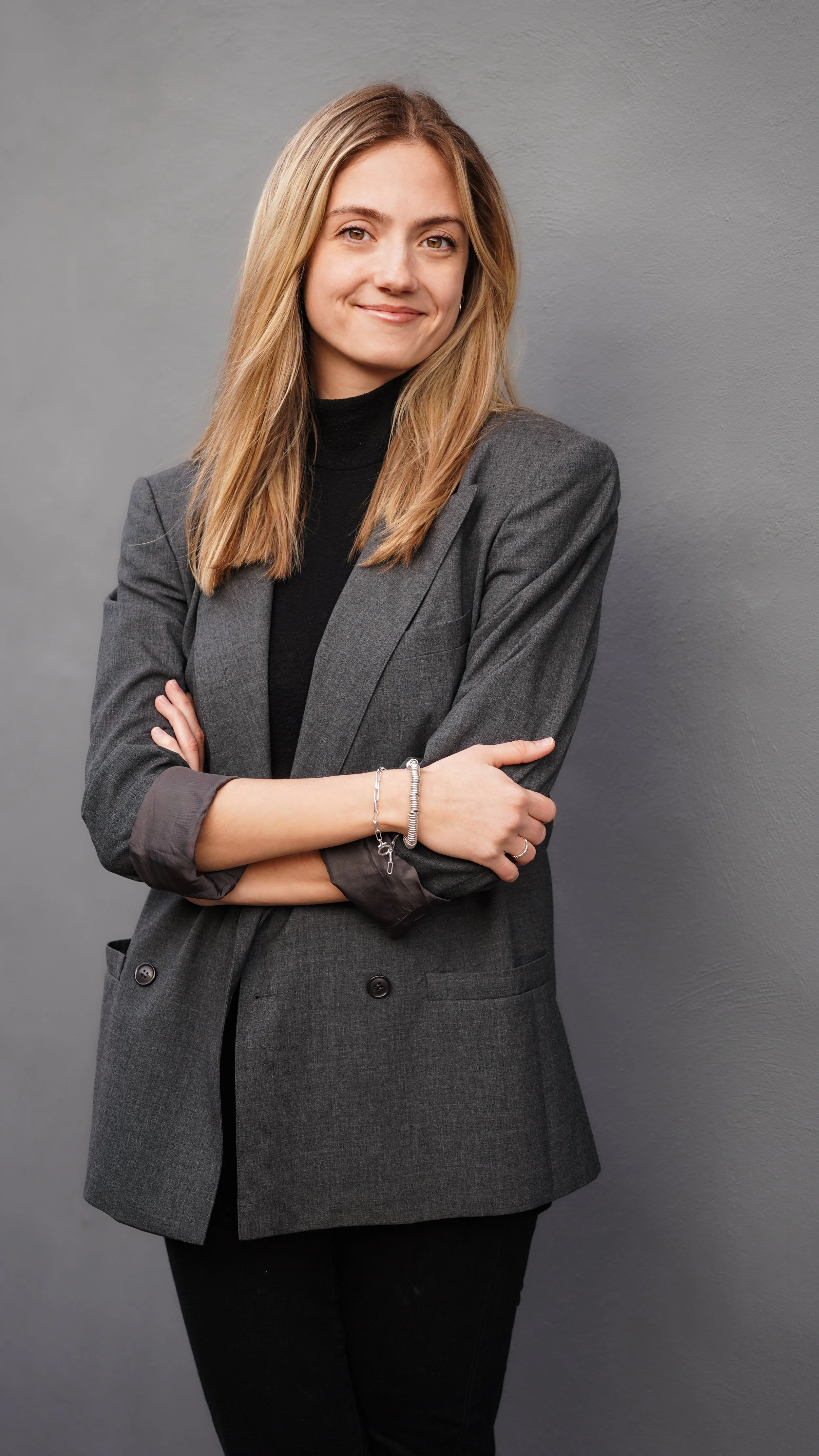 A young woman with blond hair, wearing a gray blazer over a black turtleneck, smiling and standing against a gray wall.