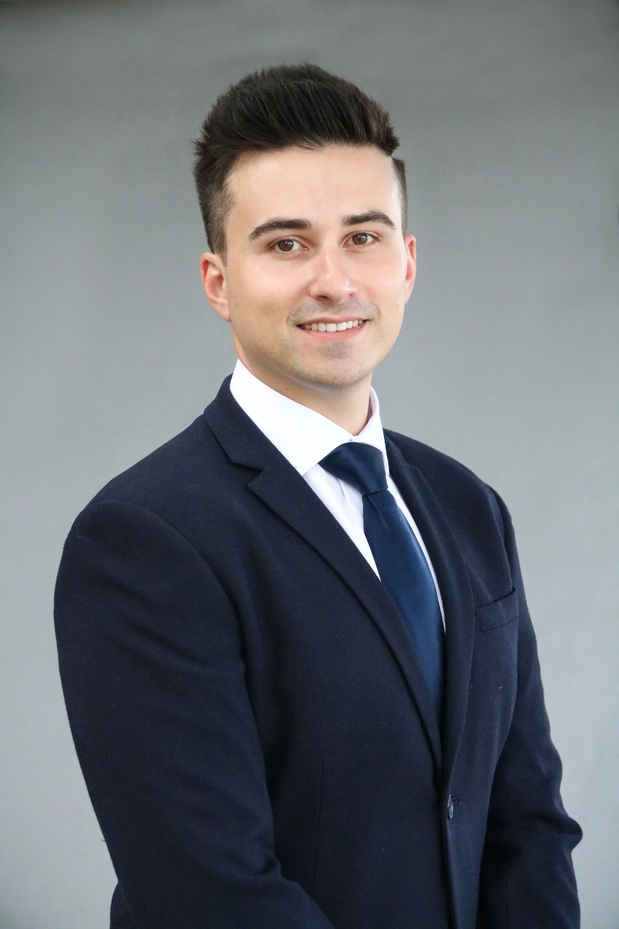A young man in a navy suit with a white shirt and a navy tie, smiling, posing against a blurred gray background.