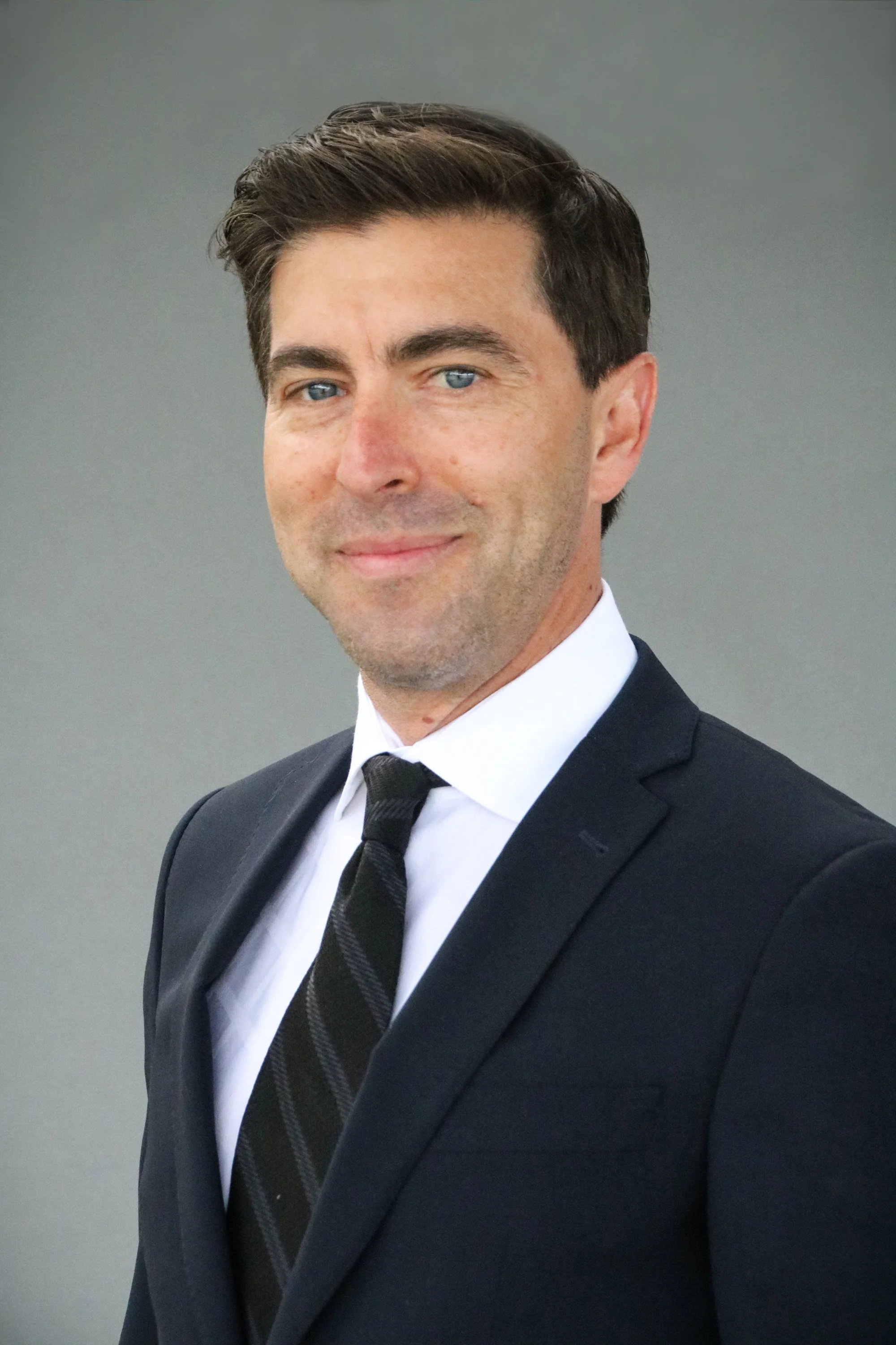 A man in a black suit, white shirt, and black tie, smiling with a neutral gray background.