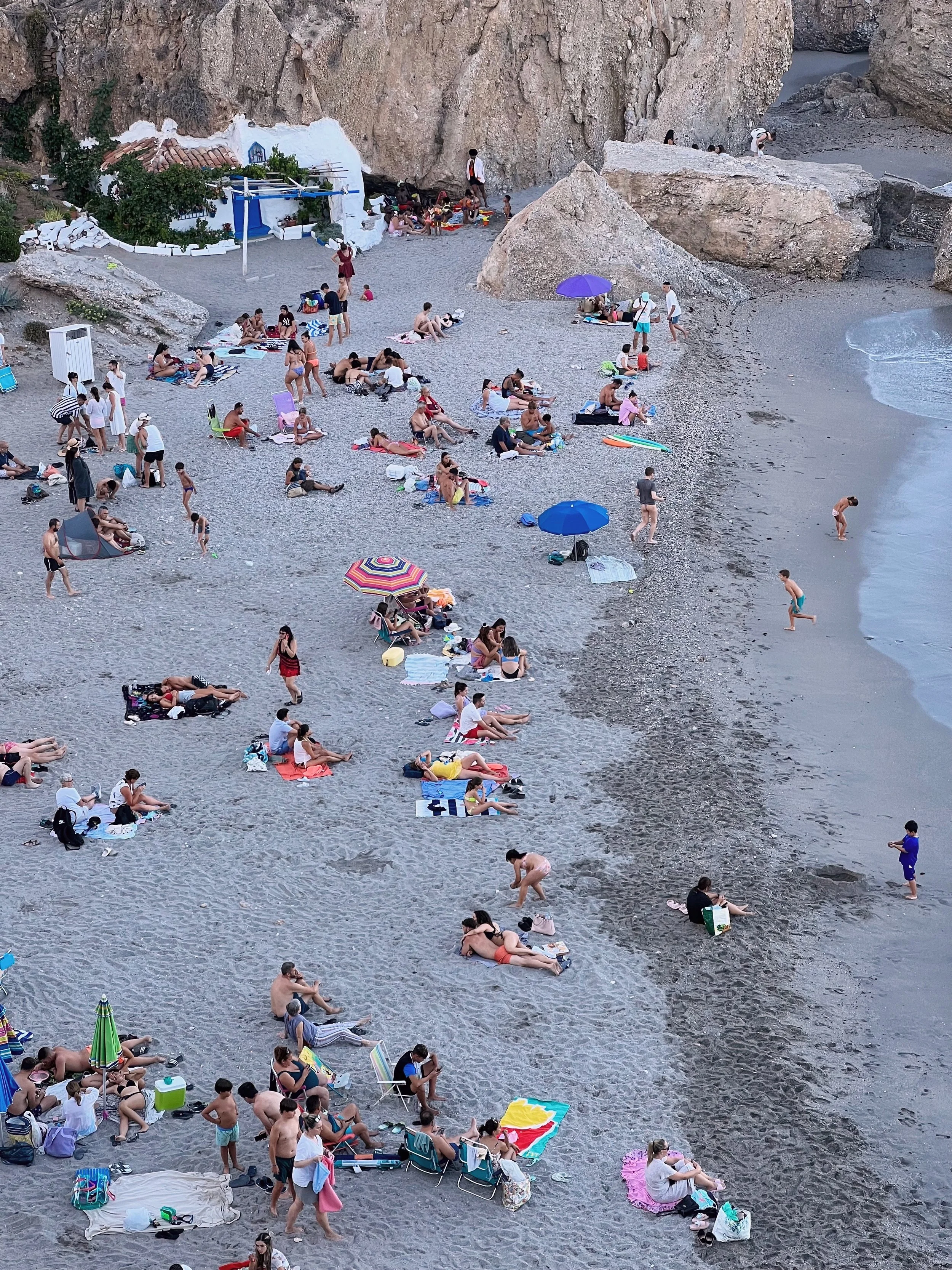 People relaxing on a beach with umbrellas, towels, and rocks, climbing areas, and a rocky backdrop.