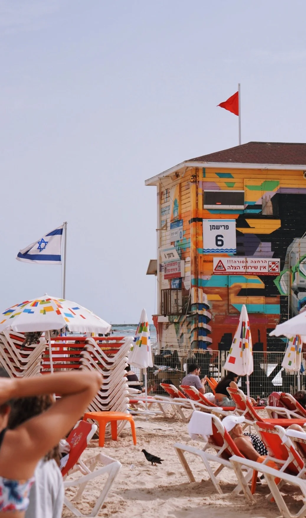 Beach scene with colorful lounge chairs and umbrellas, an Israeli flag, a graffitied building with flags on top, and people relaxing on the sand.