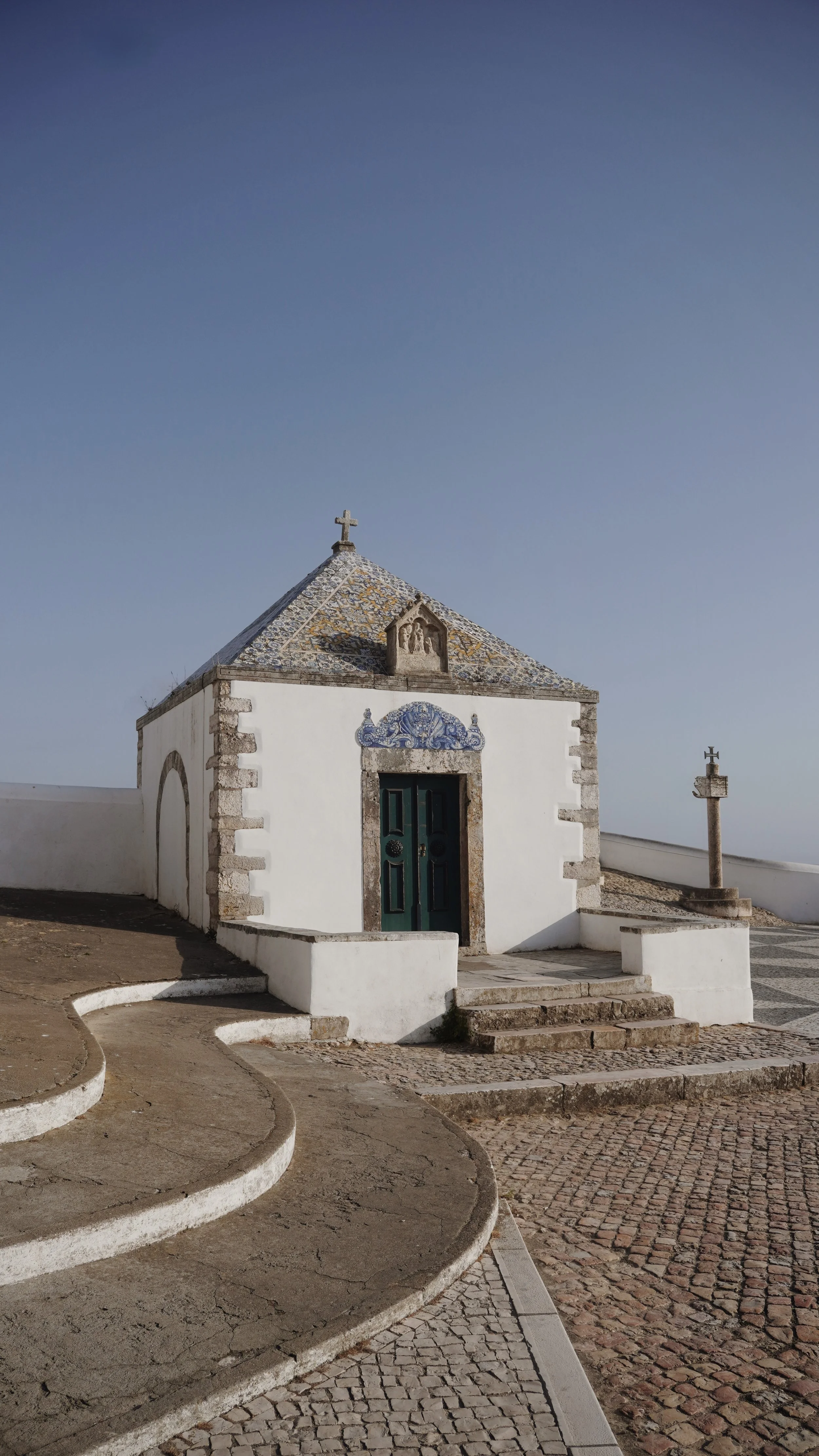 A small white chapel with a cross on the roof, stone steps leading up to a green door, and a cross monument nearby, set against a clear blue sky.