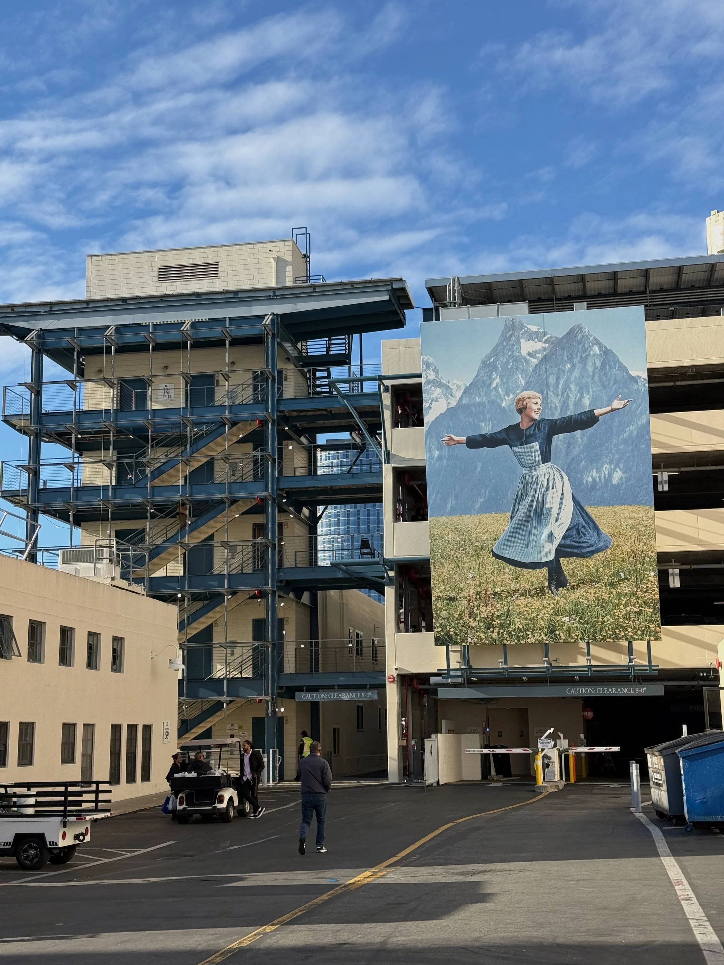 A parking garage with people and golf carts parked outside. A large mural of a woman in a traditional dress with mountains in the background is on the side of the building.