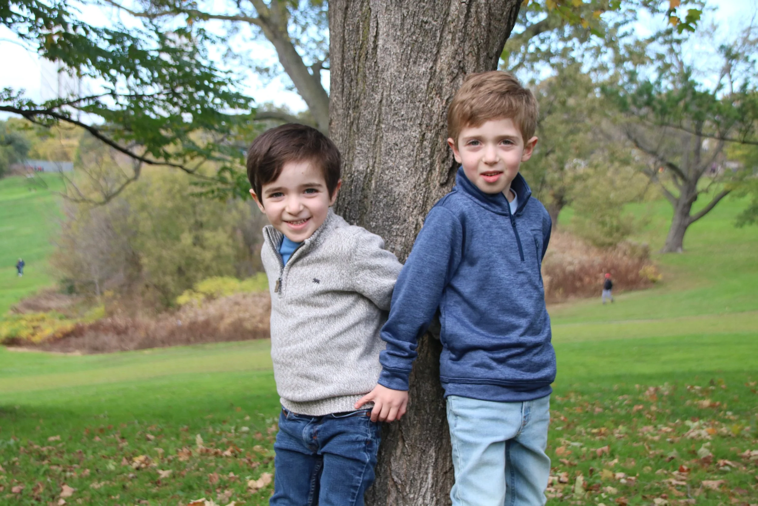 Two young boys stand back-to-back leaning against a tree in a park, smiling at the camera with grass and trees in the background.