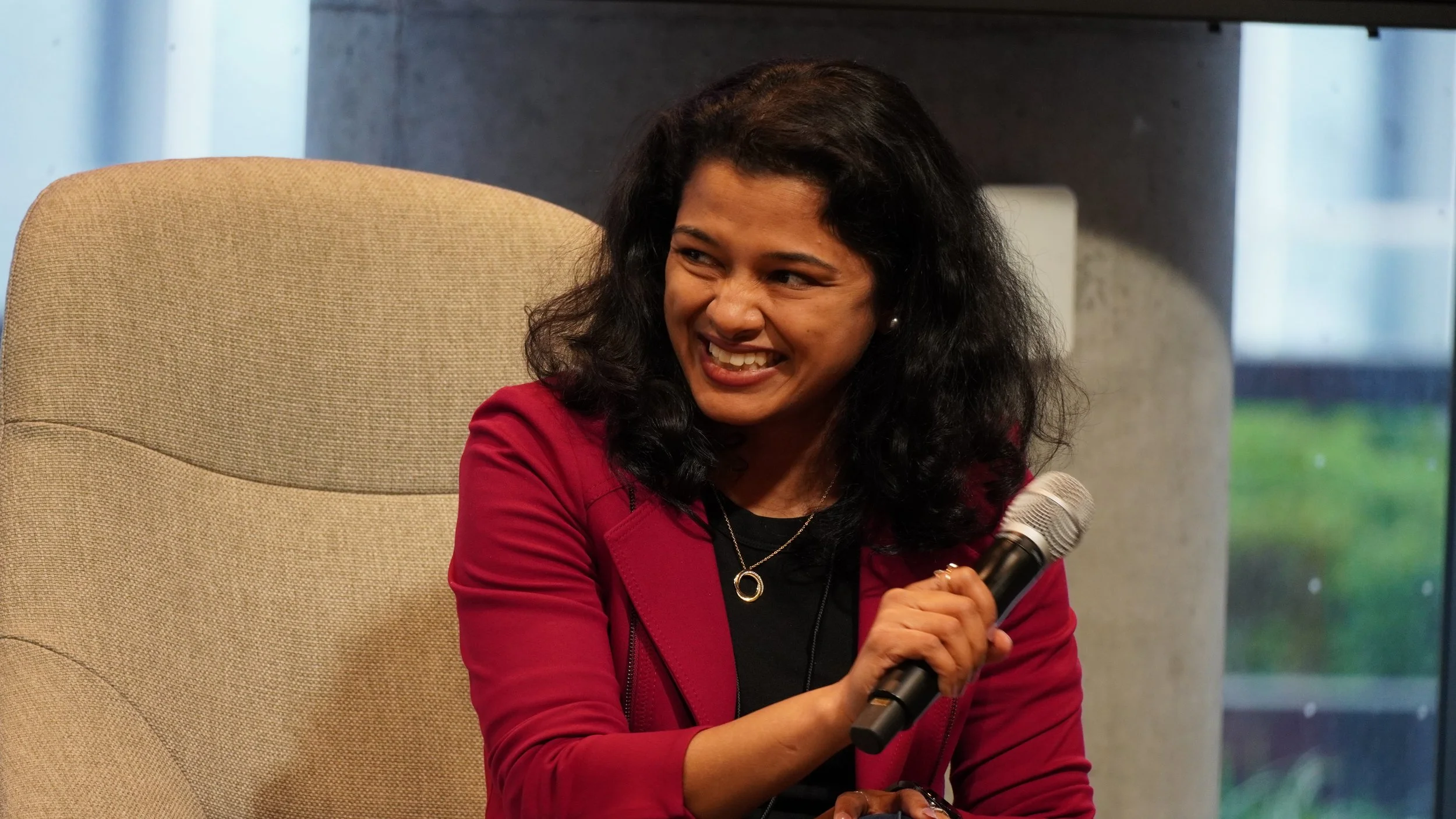 A woman with black curly hair smiling and holding a microphone, sitting in a beige chair in an indoor setting with a window showing a blurred outdoor view.