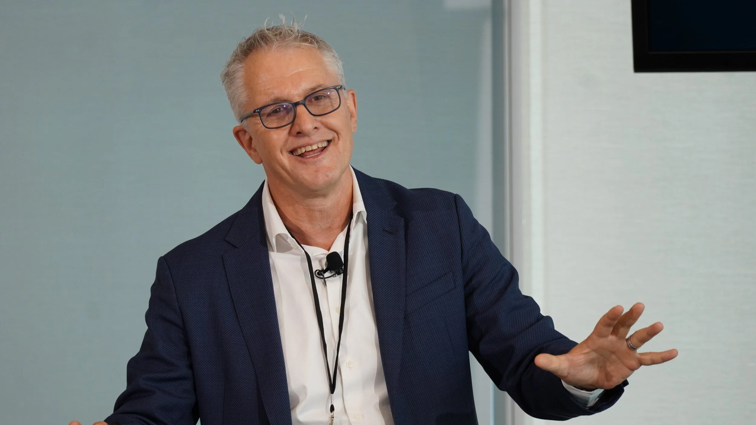A man in a dark blue blazer, white shirt, and glasses standing and smiling while gesturing with his hand during a presentation or talk.