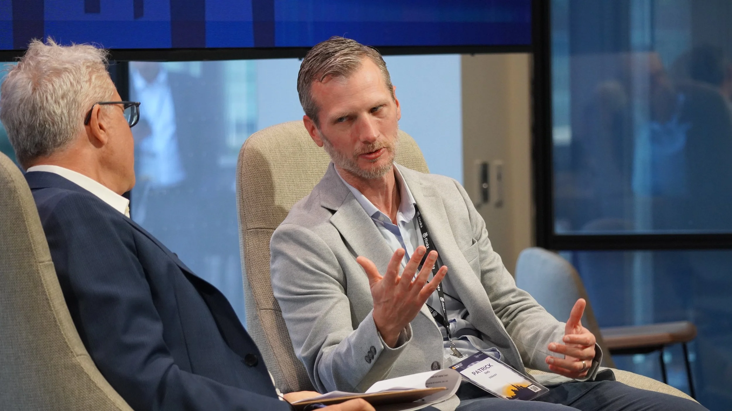 Two men are having a conversation during a conference or meeting, seated in chairs in front of a large window with city buildings visible in the background.