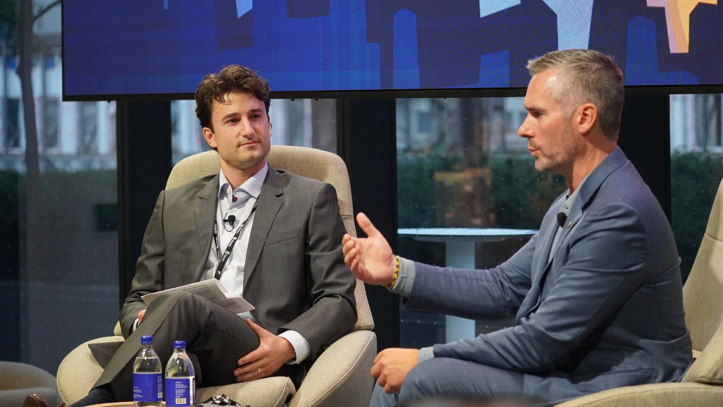 Two men in business suits engaged in conversation while seated in chairs during an event or interview, with a large digital screen behind them displaying blue graphics.