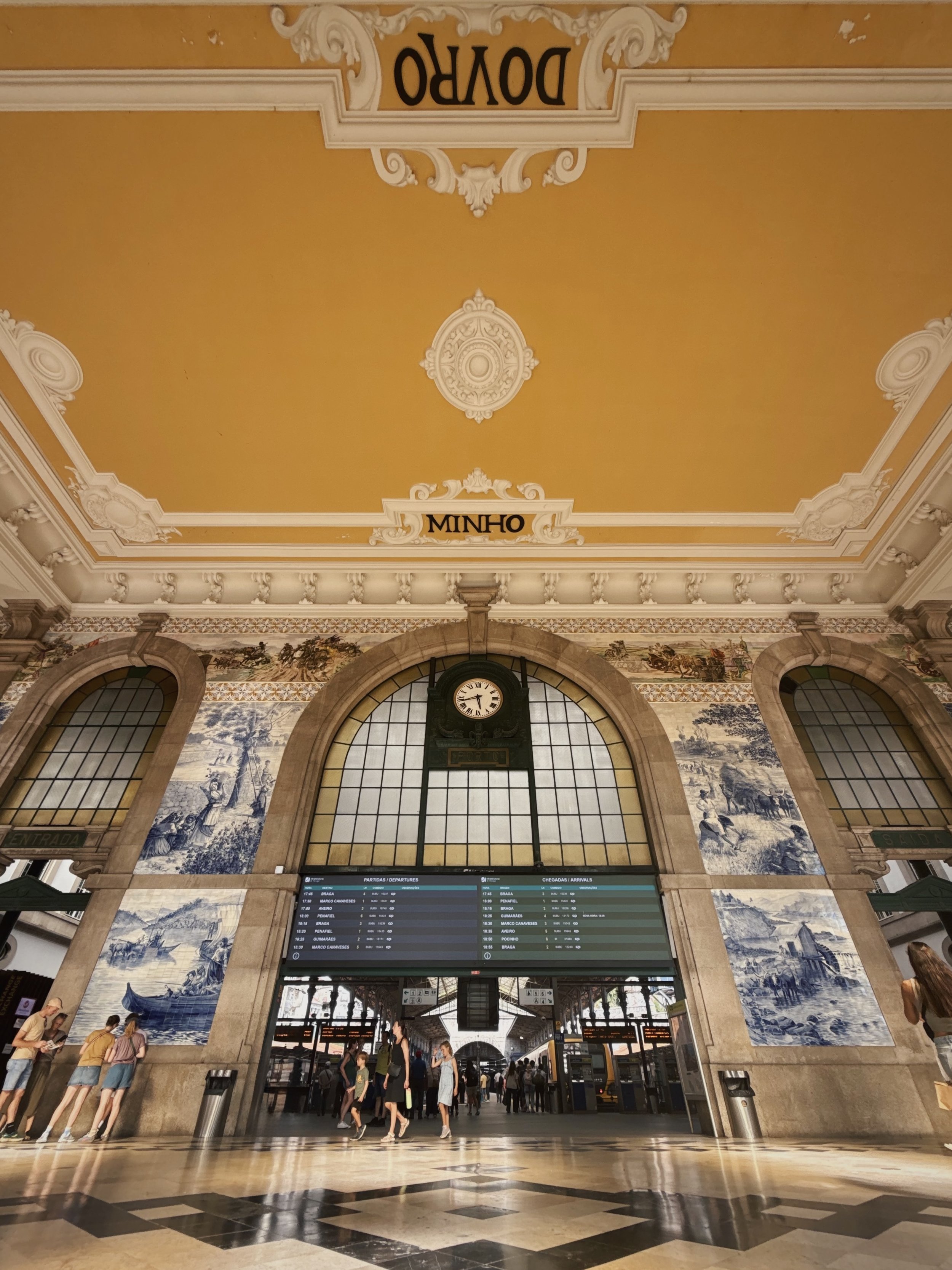 Tourists walking inside a historic train station with a large clock, decorative blue and white tiled murals, large arched windows, and an electronic departure and arrival board.