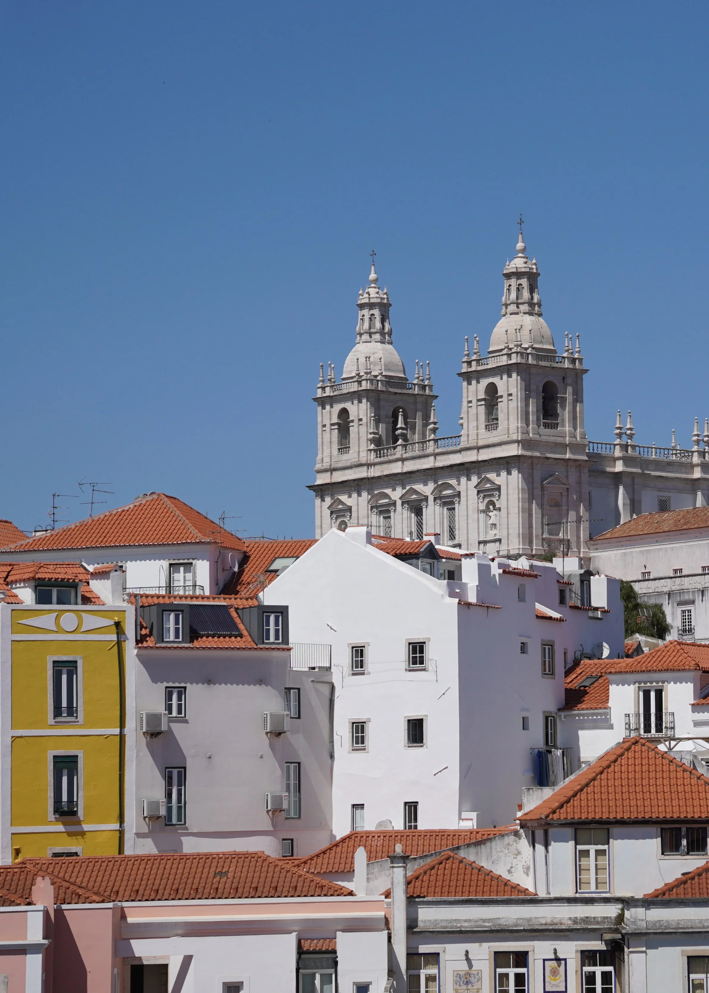 White buildings with red-tiled roofs and a large historic church with two towers in the background under a clear blue sky.