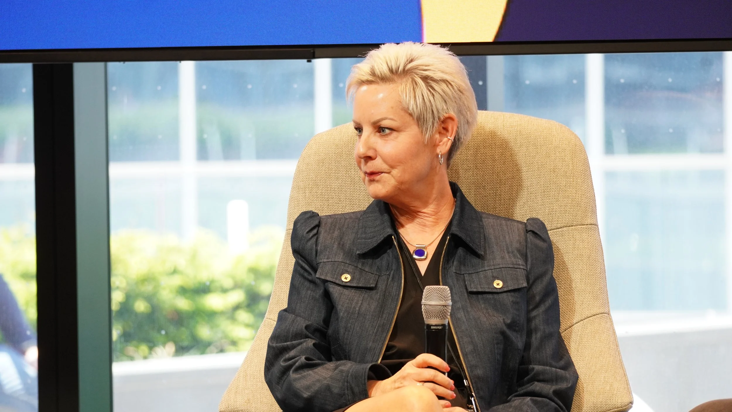A woman with short gray hair, wearing a black jacket, sitting in a beige chair holding a microphone, looking to her right, with a large window and cityscape in the background.