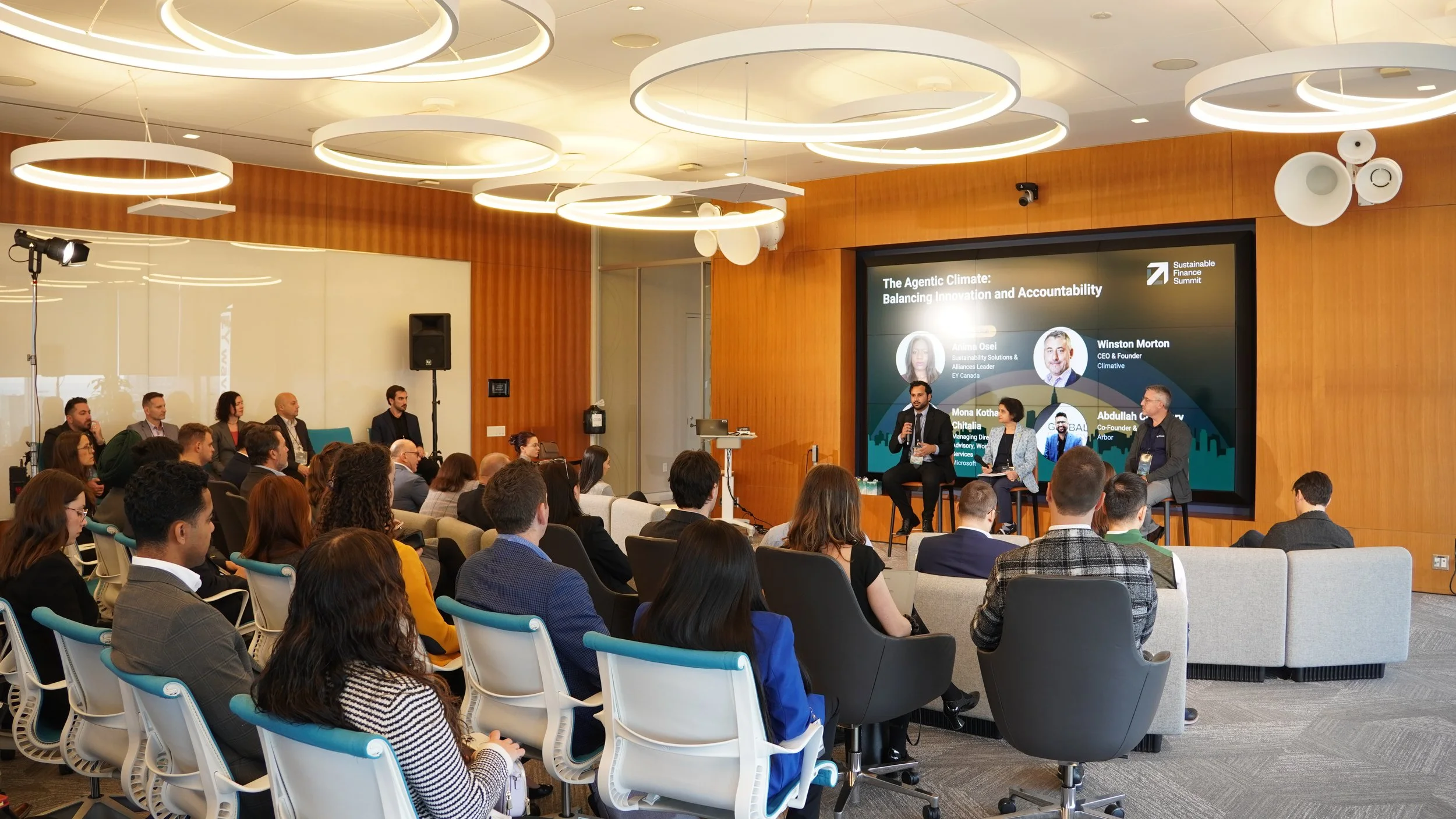 A panel discussion at a conference with four speakers on stage and an audience seated in a modern conference room. The screen behind the speakers displays titles and participant names.