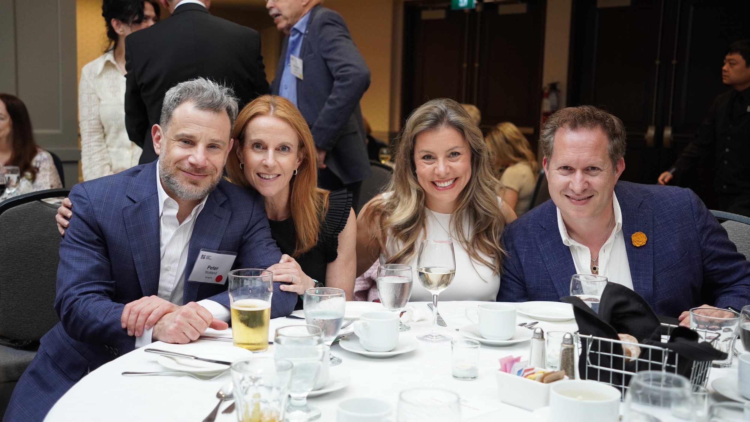 Four people sitting at a dinner table, smiling, with drinks and tableware, in a formal event setting.