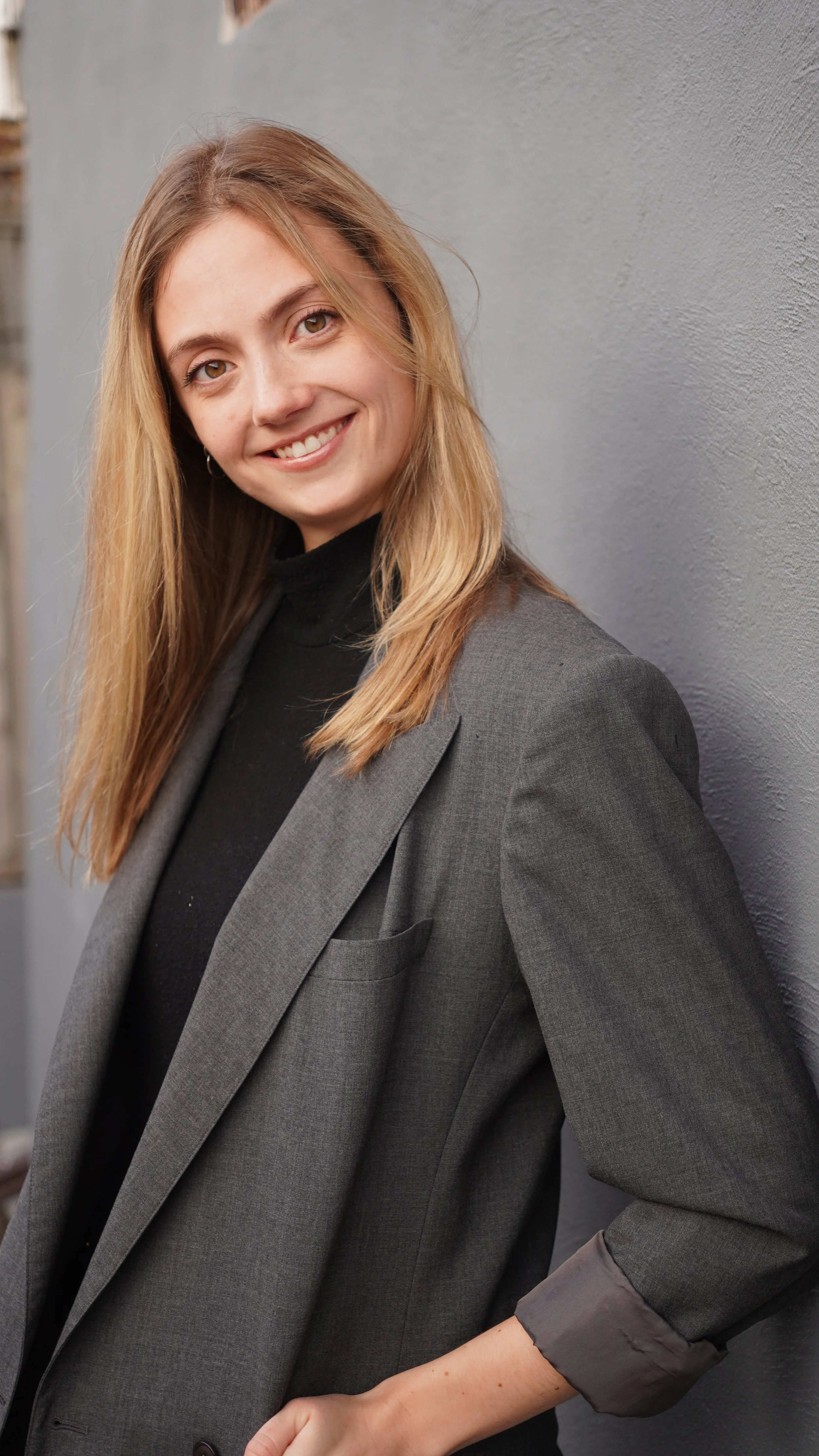 A woman with long blonde hair smiles while leaning against a gray wall, wearing a dark gray blazer over a black turtleneck.