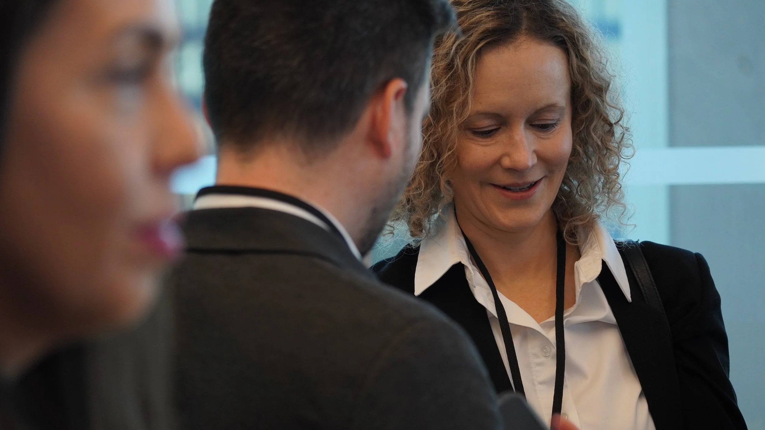 Businesspeople in a meeting, focusing on two women, one with curly hair smiling and a man with short dark hair, all dressed in professional attire.