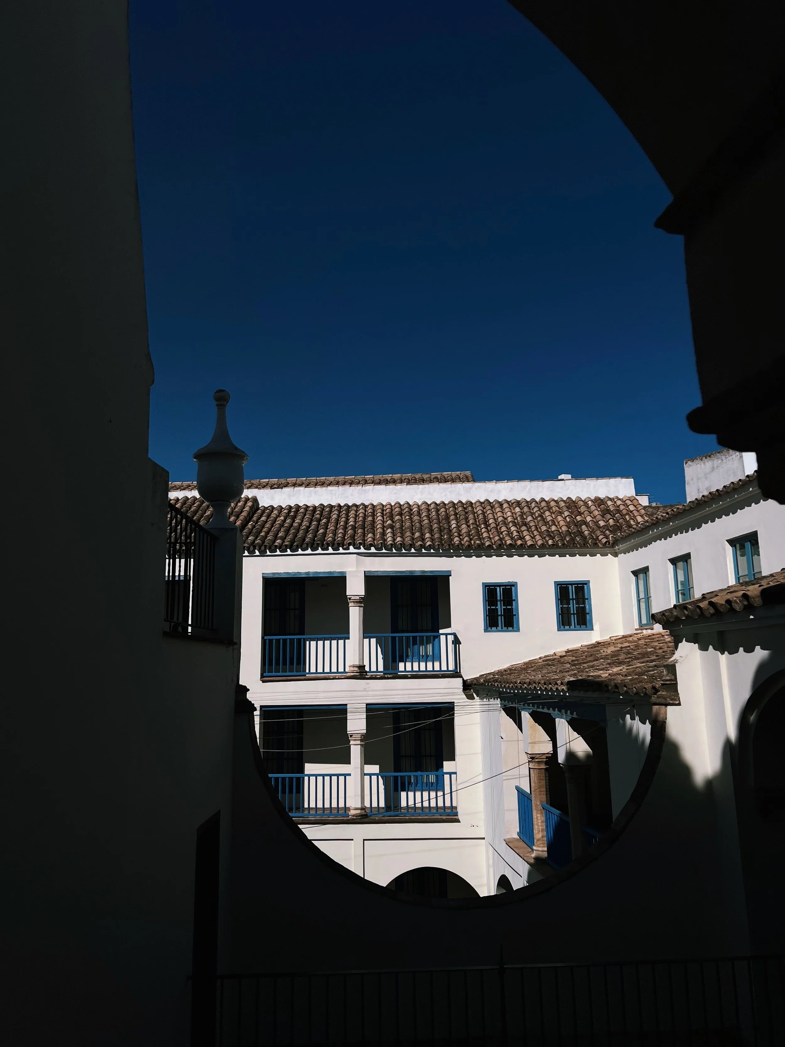 View of a white multi-story building with blue railings and windows, and a terracotta tiled roof, framed by dark shadows from surrounding walls, under a clear dark blue sky.