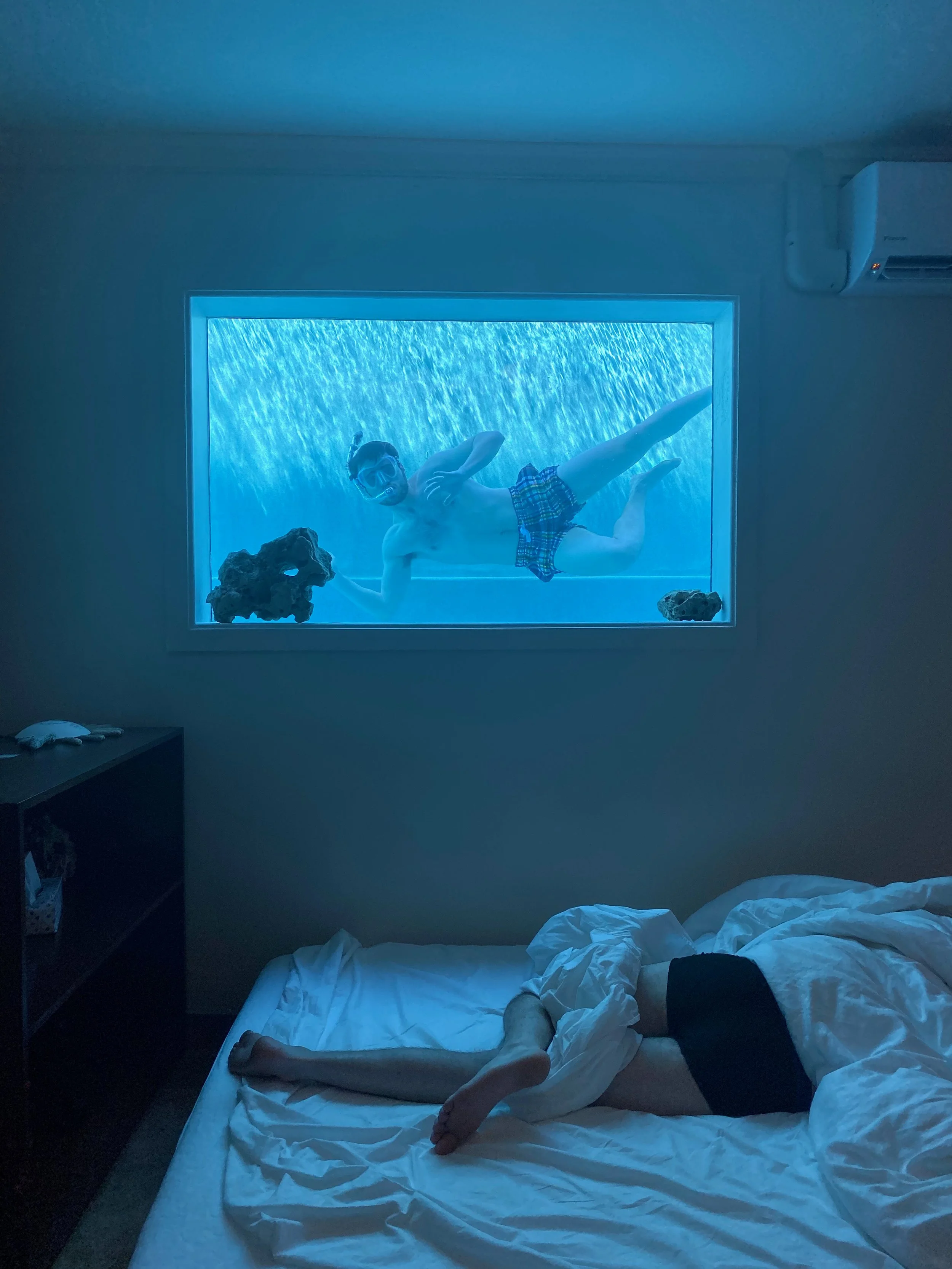 A person lying on a bed with white sheets, partially covered, with legs visible. Outside a window above the bed, a man in swim trunks and goggles is floating in a pool, making a peace sign with his hand, with a waterfall or water feature in the backg