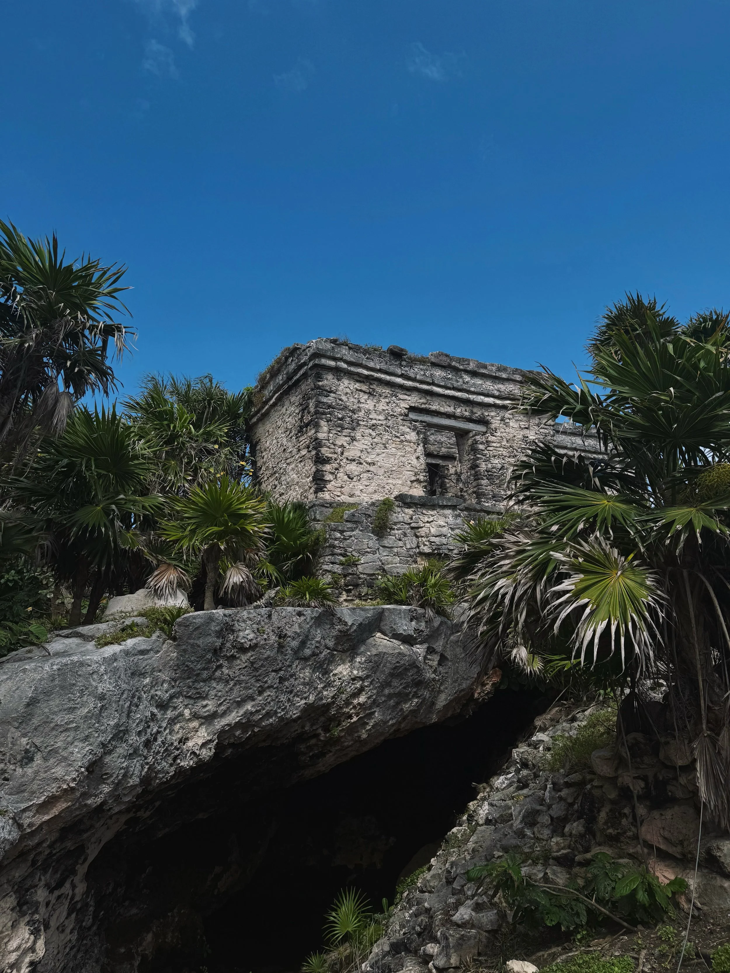 Ancient stone Mayan ruin atop a rocky hill, surrounded by lush green tropical plants, against a clear blue sky.