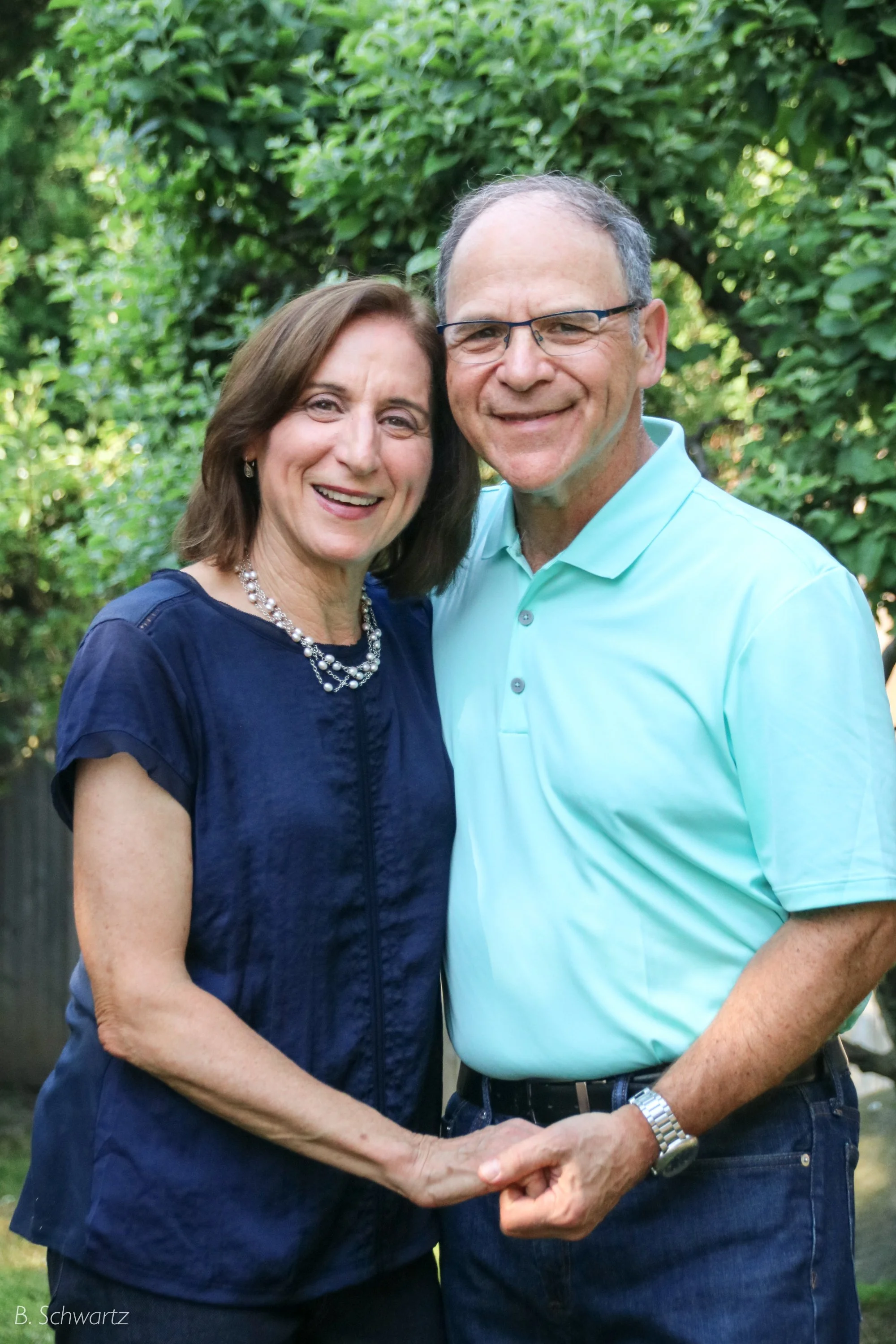 A smiling middle-aged couple standing close together outdoors, holding hands, surrounded by green trees.