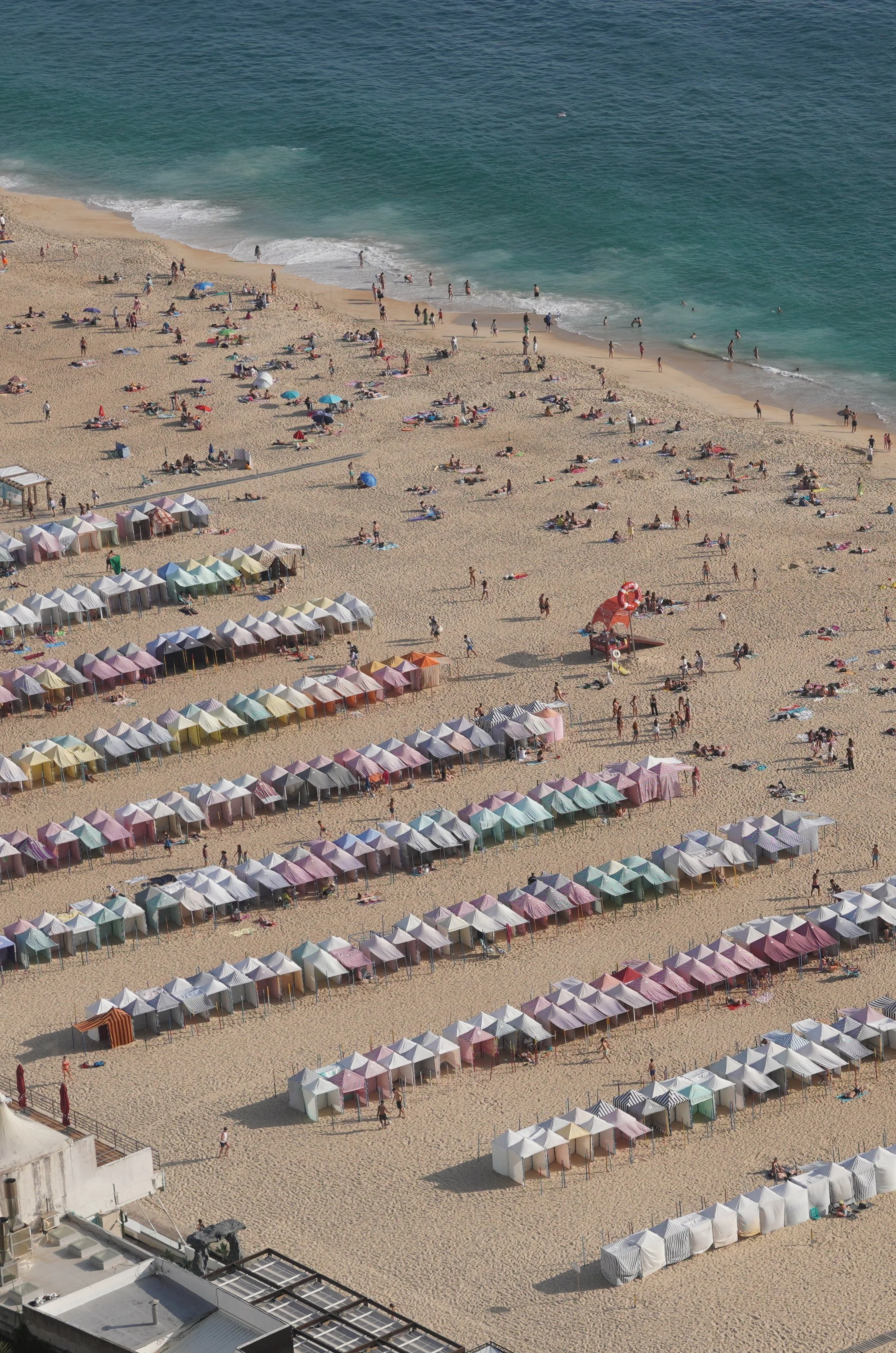 A crowded beach with colorful tents in rows, sunbathers, and people near the shoreline with the ocean in the background.