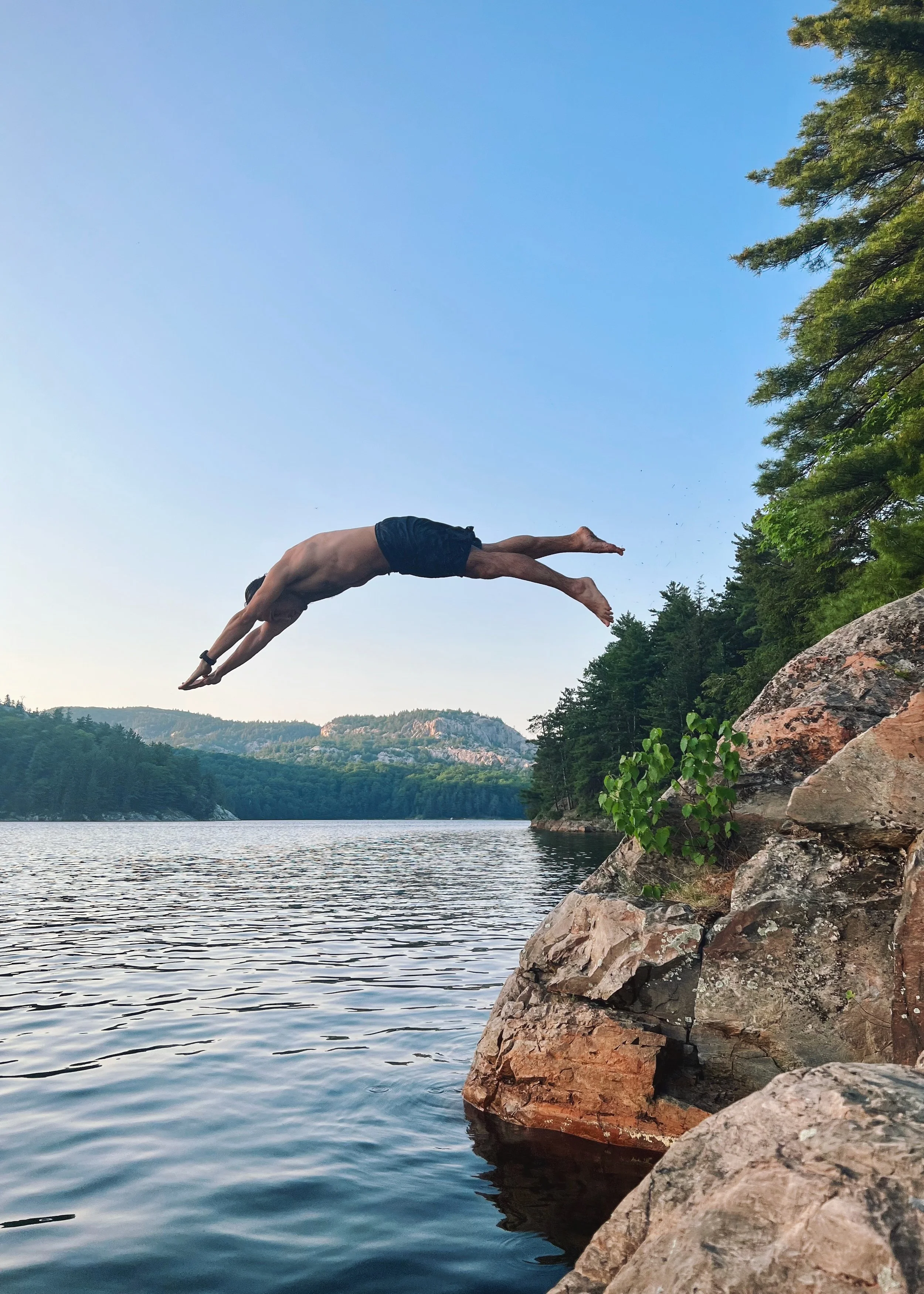 A man diving from a rocky cliff into a lake with forested hills in the background under a clear sky.