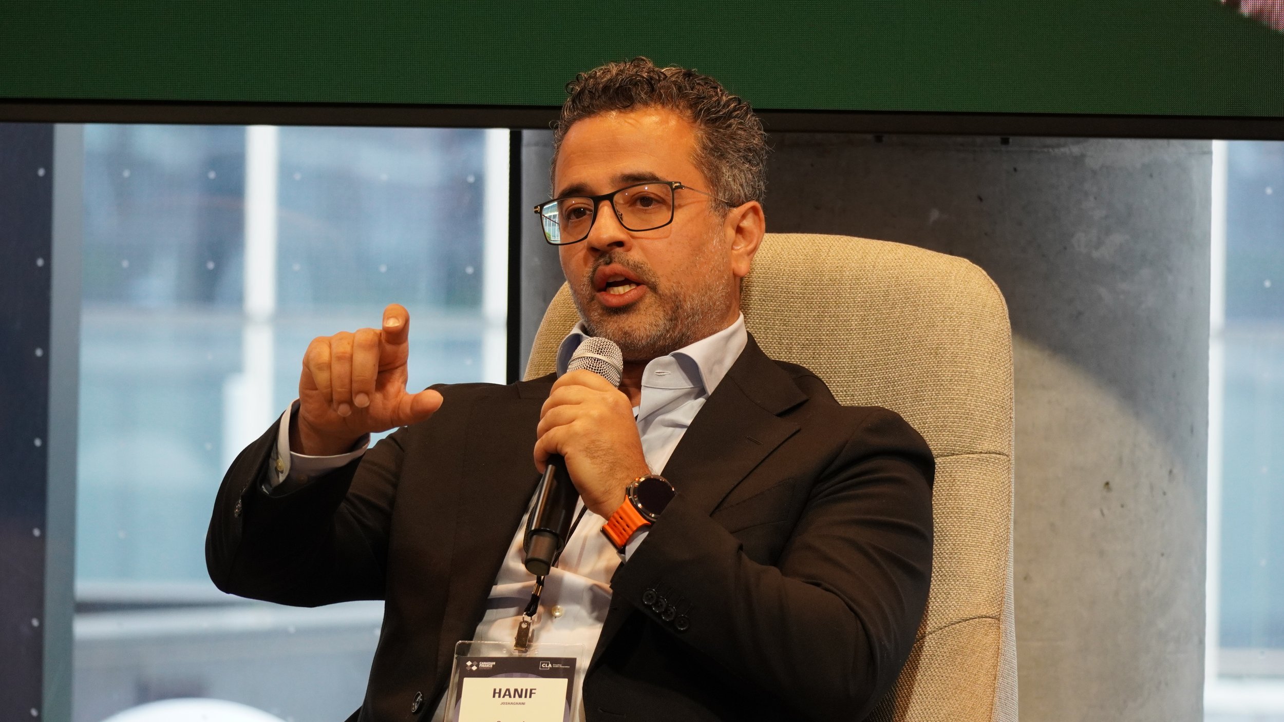 A man with glasses and a beard, wearing a black suit and white shirt, is sitting in a beige chair, speaking into a microphone during a conference or interview, with a name badge that says 'HANIF' around his neck.