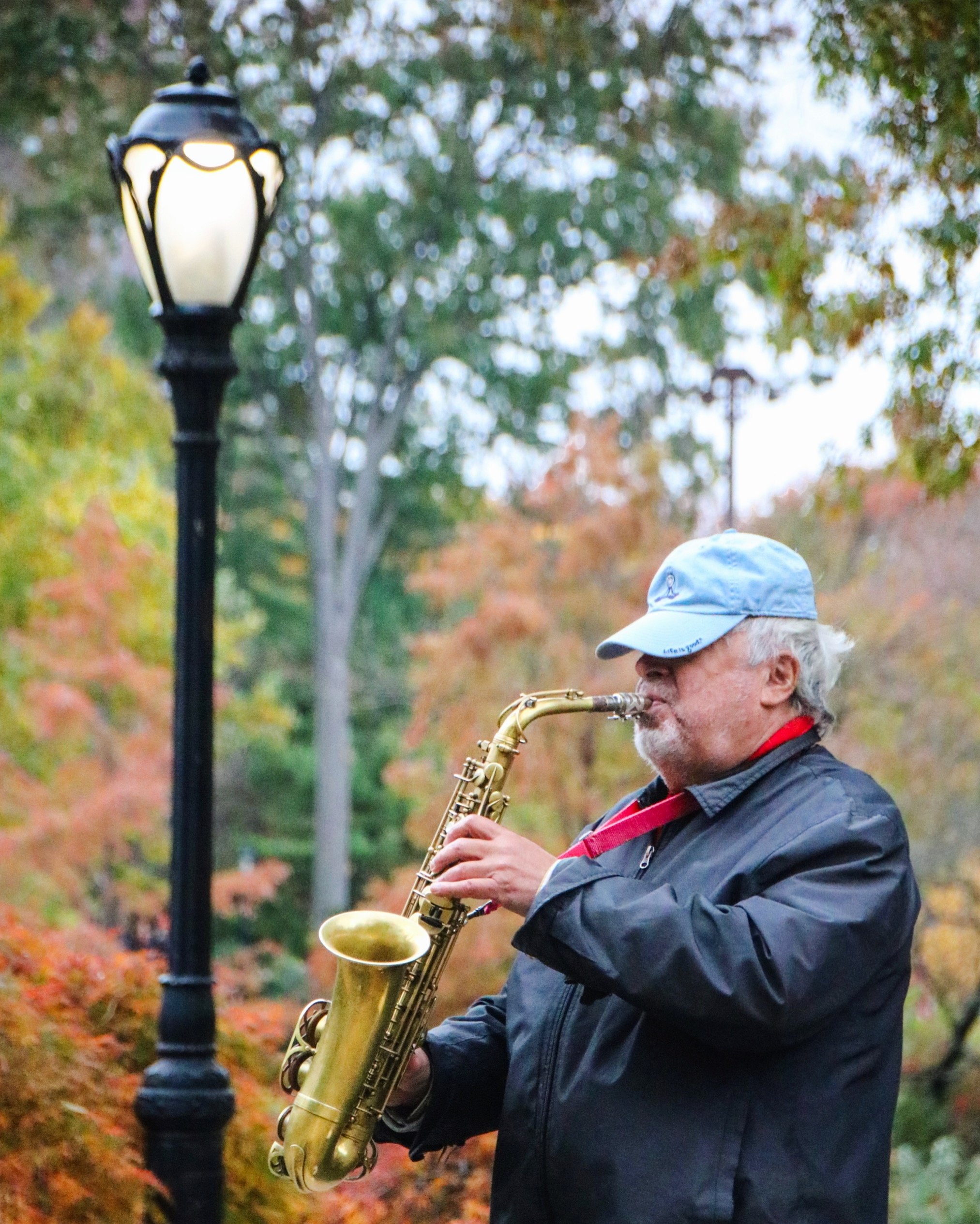 Older man playing saxophone outdoors during autumn, wearing a blue cap and black jacket, with fall foliage trees in the background and a streetlamp nearby.