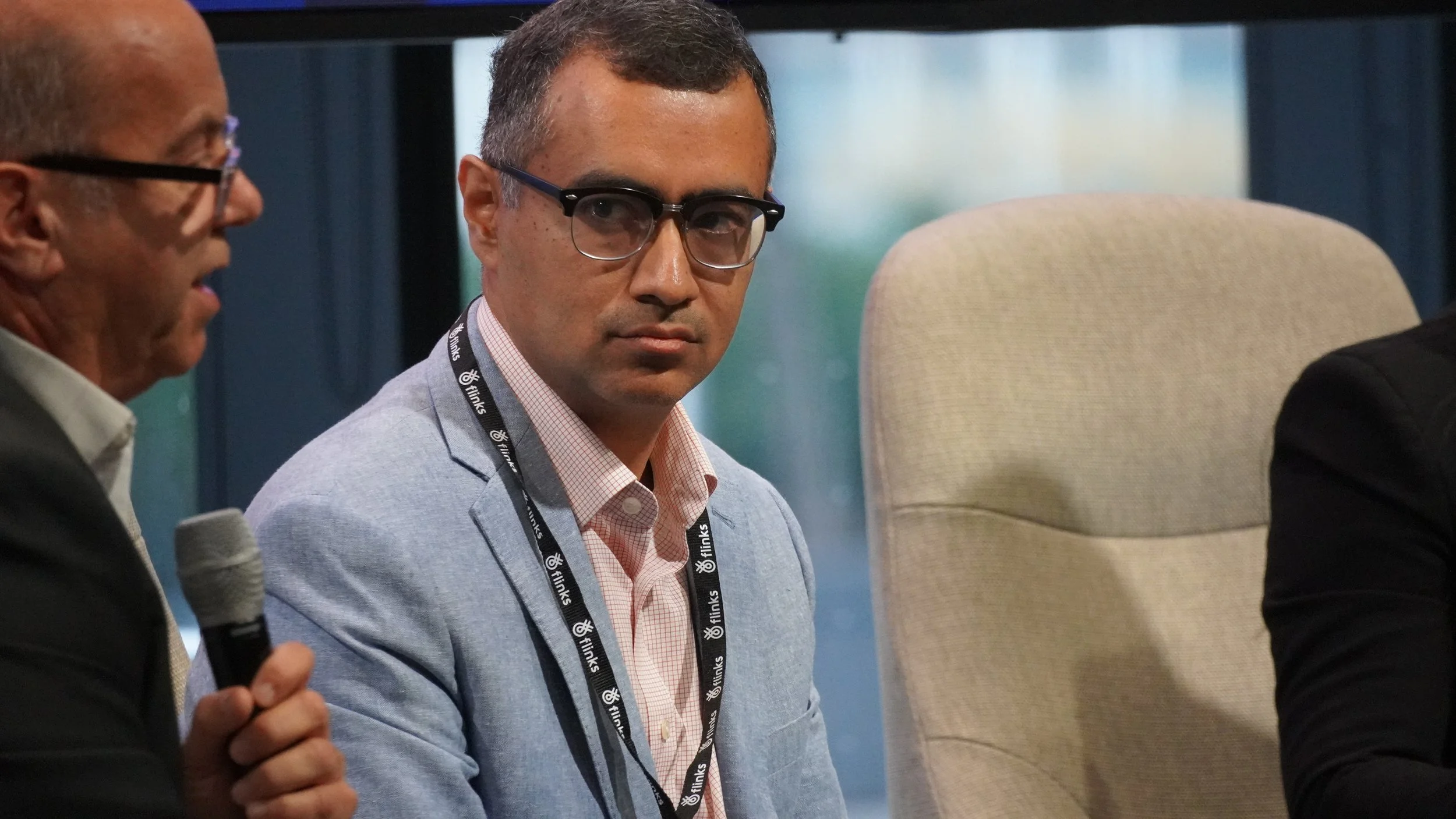 A man in glasses wearing a light blue blazer and pink shirt is sitting next to a beige chair, during a discussion or meeting, with another man holding a microphone partially visible on the left.