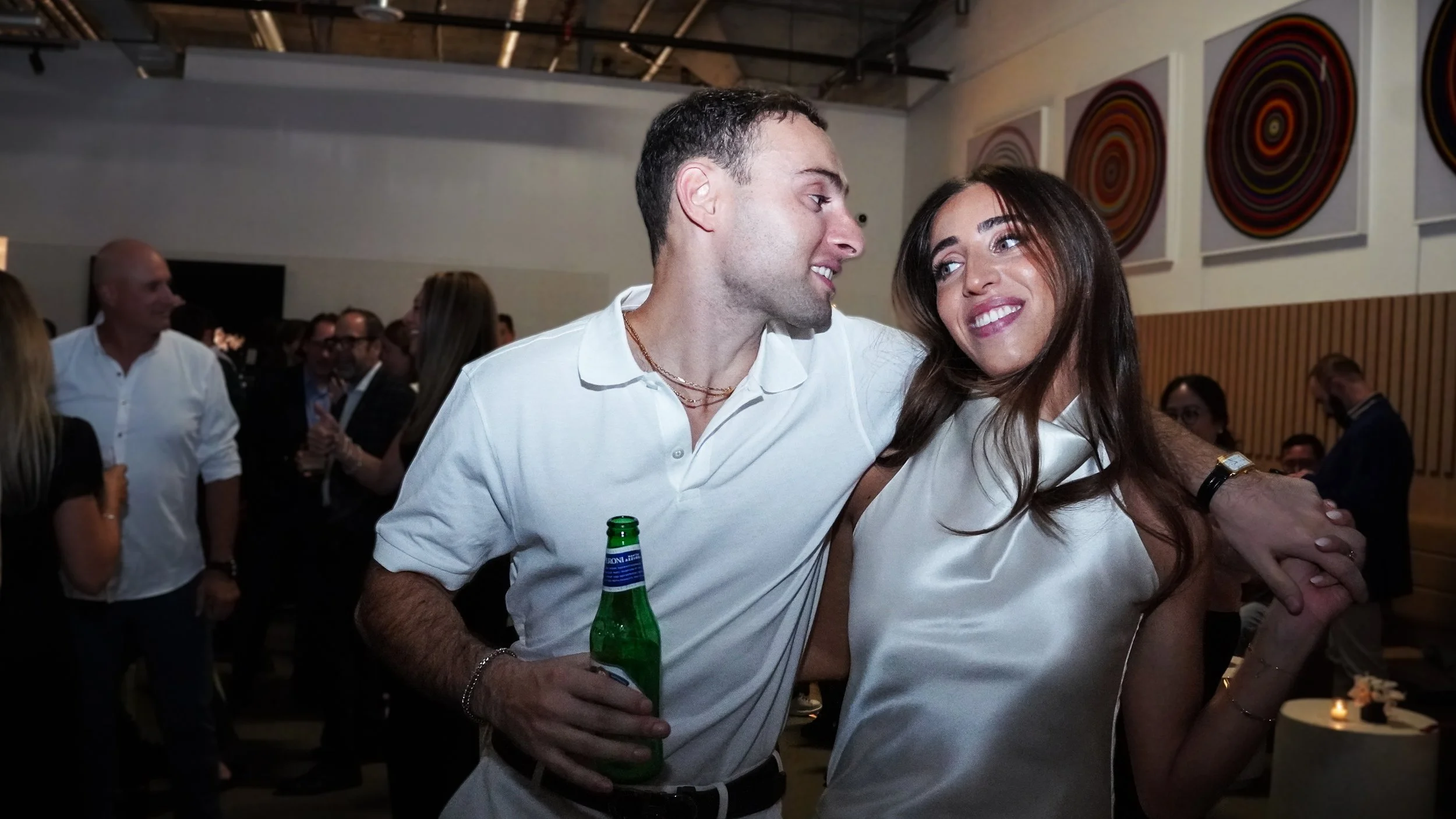 A man with short dark hair wearing a white polo shirt, holding a green bottle, and a woman with long brown hair wearing a white satin top, smiling and looking at each other at a social gathering with people in the background and colorful circular wal